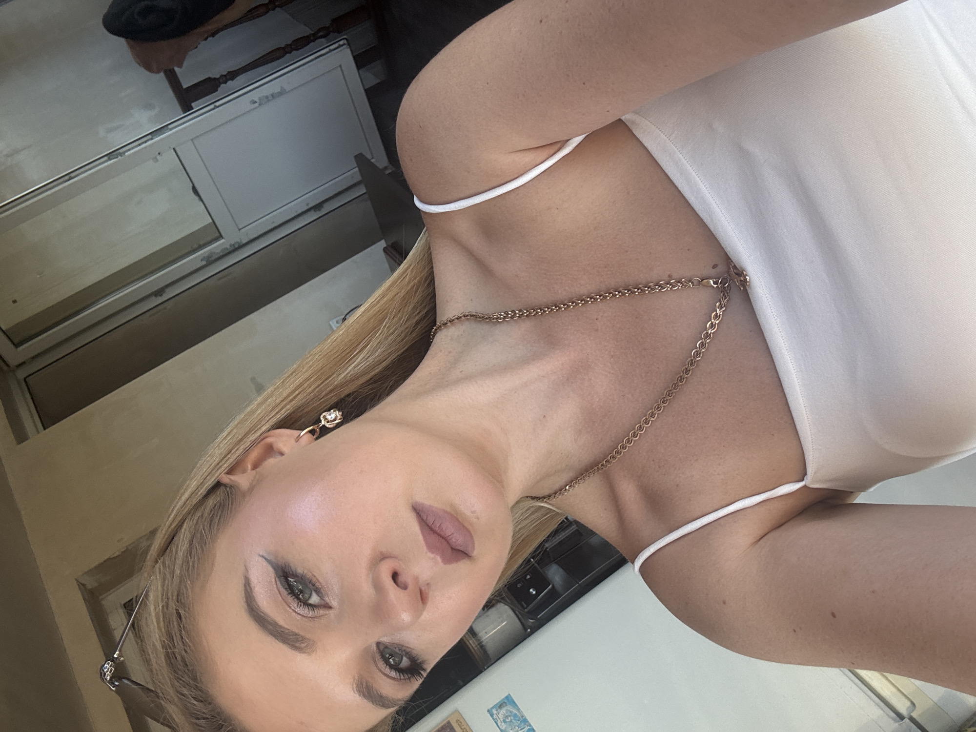 The image is a close-up, vertically oriented photograph of a young woman. She has blonde hair and is wearing makeup, including eyeliner and lipstick. She is wearing a white tank top with thin straps and a gold chain necklace. She is also wearing earrings. The background is blurred and appears to be indoors, possibly in a home or studio setting, with hints of furniture and possibly a window. The lighting suggests it could be daytime. The woman's expression is neutral. There is no visible text in the image.