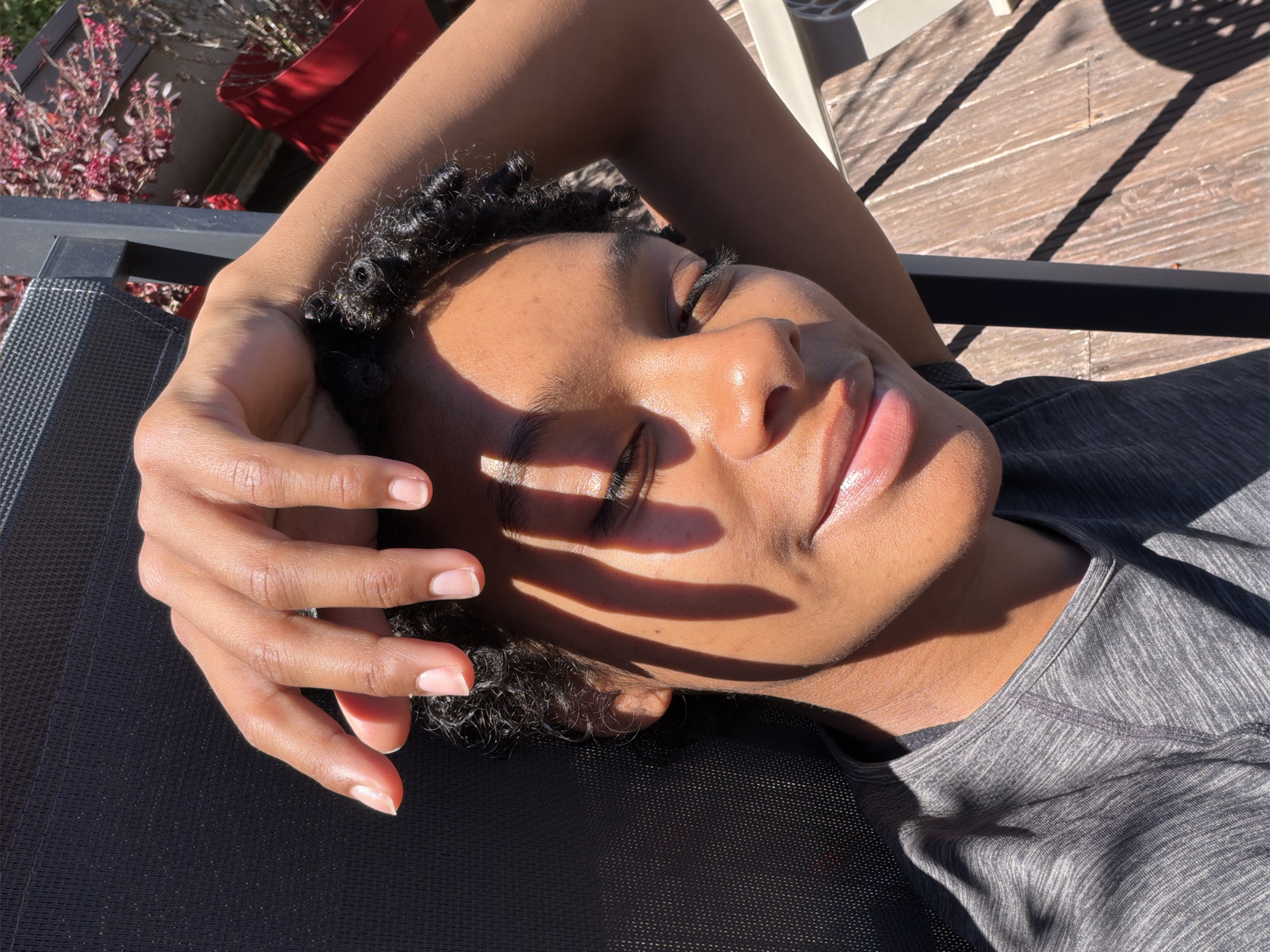 A medium-shot of a Black woman with curly hair is laying down outdoors on a black lounger with mesh webbing. Her eyes are closed, and she has one arm raised, with her hand resting on her forehead, casting shadows on her face. She is wearing a grey heathered shirt. The lounger is placed on a wooden deck. The background features plants in a red container. There are hints of a white chair. The location is the city of Champs-sur-Marne, France.
