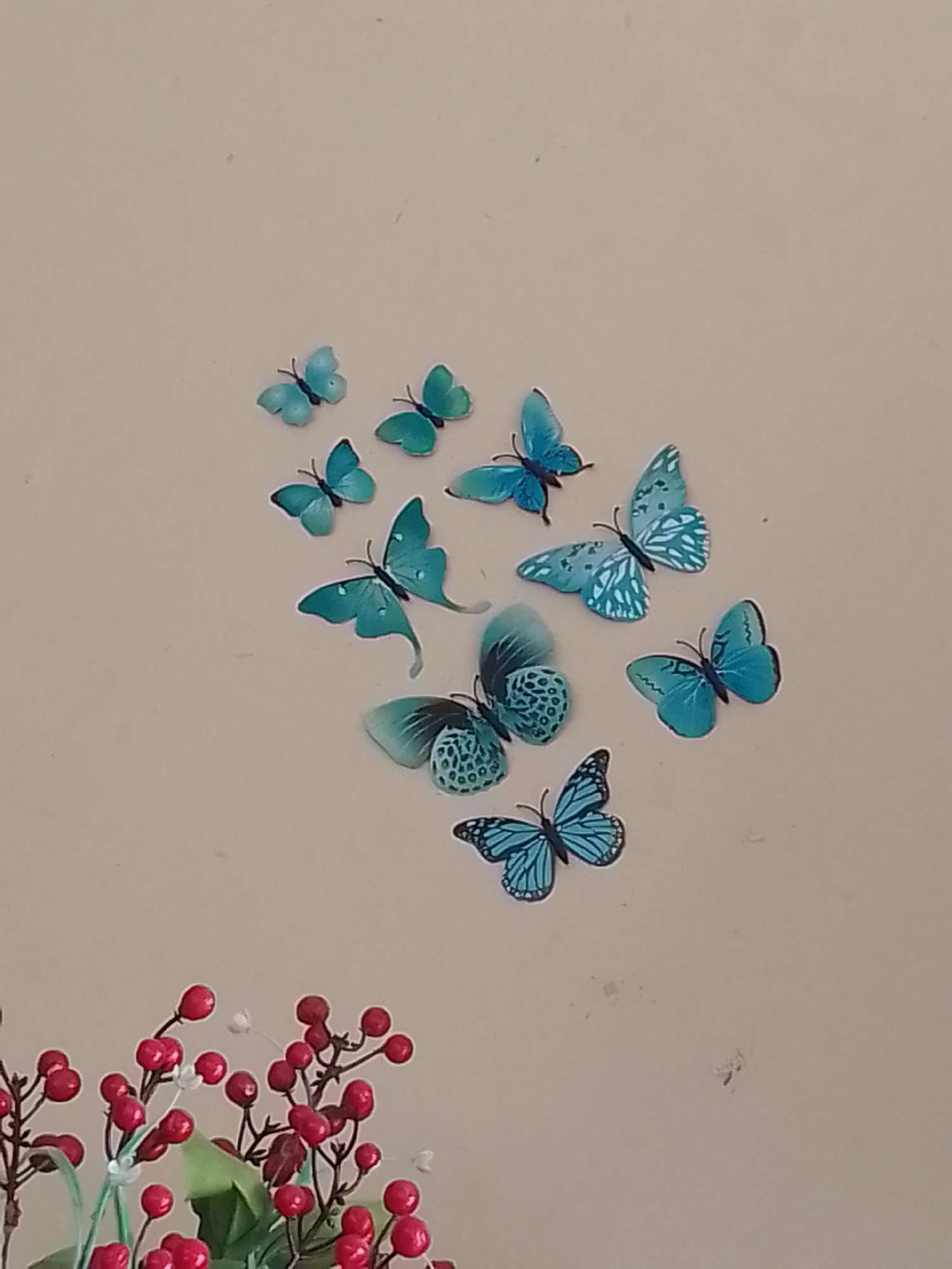 The image shows a collection of decorative butterflies on a plain, beige wall. The butterflies are predominantly blue and turquoise, with varying patterns and sizes, and are arranged in a scattered pattern across the upper right portion of the wall. In the lower left corner of the image, a cluster of red berries on thin stems, along with some green foliage, is visible. This arrangement suggests a decorative display, possibly for a room or event. The image does not contain any information that would allow for its location to be identified as being in the city of Bukittinggi, Indonesia.