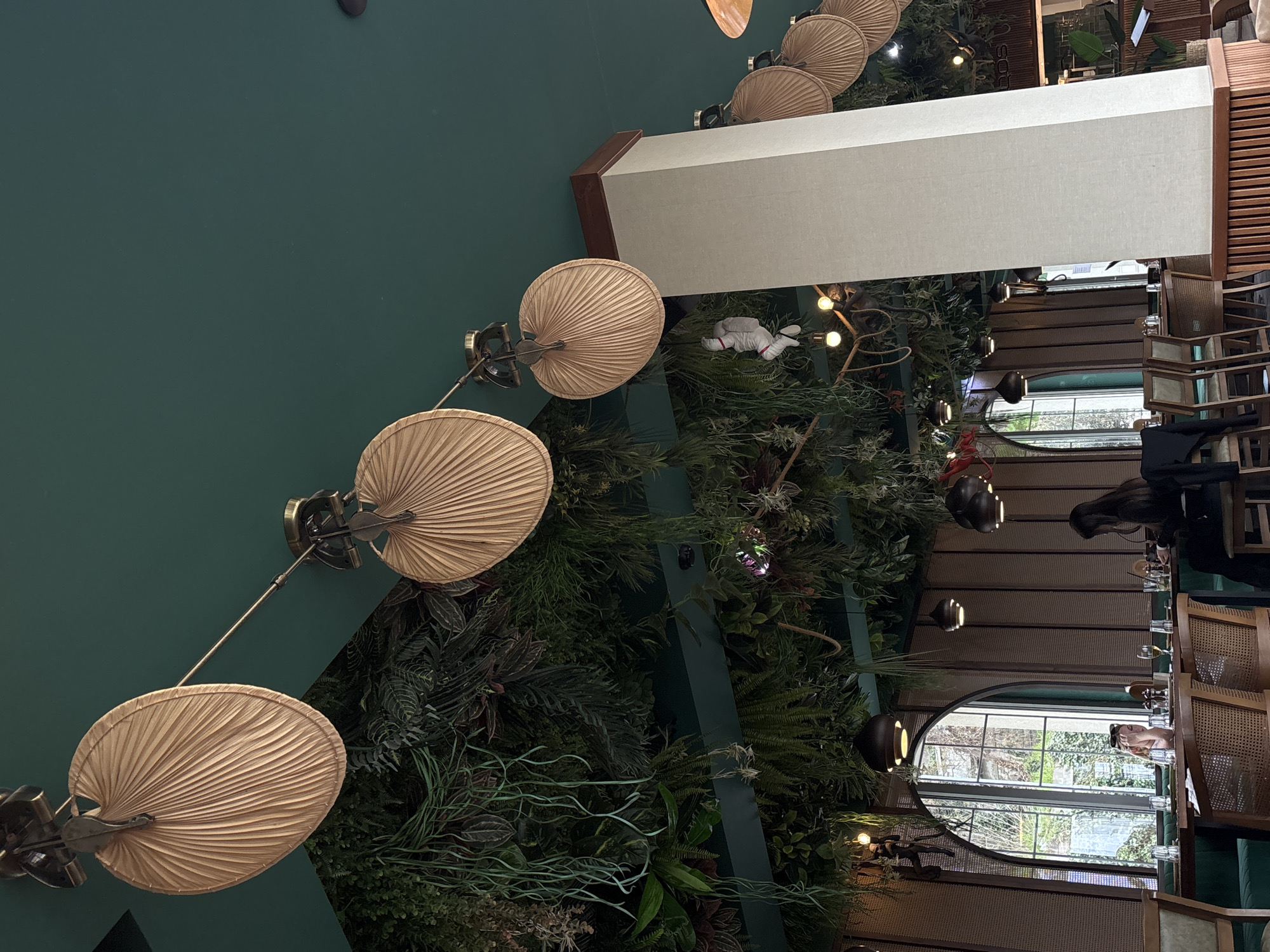 Captured from an unusual angle, the photograph presents an interior shot featuring a ceiling adorned with multiple fan-like fixtures extending downward, set against a backdrop of dense greenery. This vegetative arrangement, comprised of various plants and foliage, is situated near what appears to be a dining area. The dining space includes chairs, tables set with glassware, and visible window frames suggesting an exterior view. People are seated at the tables, although their specific actions are not readily discernible from the image.
Given the description of Varna, Bulgaria, the setting implies an establishment with a decor that integrates nature and contemporary design elements.