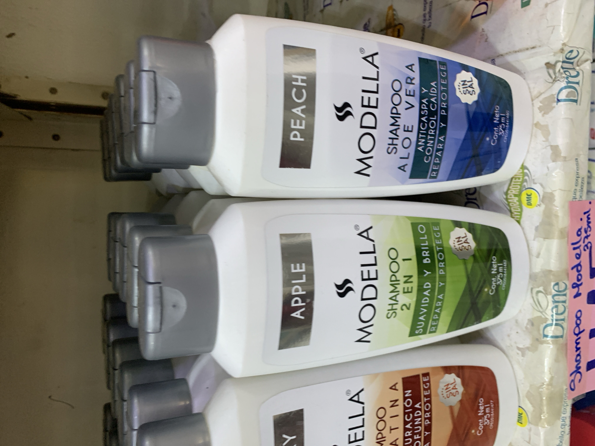 The image shows a close-up of several bottles of shampoo lined up on a shelf. The bottles are predominantly white with silver caps. The labels on the bottles are in Spanish. Two prominent labels are visible: one for "APPLE" shampoo and another for "PEACH" shampoo, both from the brand "MODELLA". Below these, other text indicates "SHAMPOO 2 EN 1" and claims of "SUAVIDAD Y BRILLO" (Softness and Shine) and "REPARA Y PROTEGE" (Repairs and Protects). Another bottle to the left has a label that is partially obscured but appears to say "KERATINA" (Keratin). A label at the bottom of each visible bottle indicates "Cont. Neto 375 ml" (Net Content 375 ml). The background is slightly blurred, showing more rows of similar products. The overall setting appears to be a retail environment, likely a store or supermarket shelf. There is no information within the image to ascertain the location as Santa Ana, Venezuela.