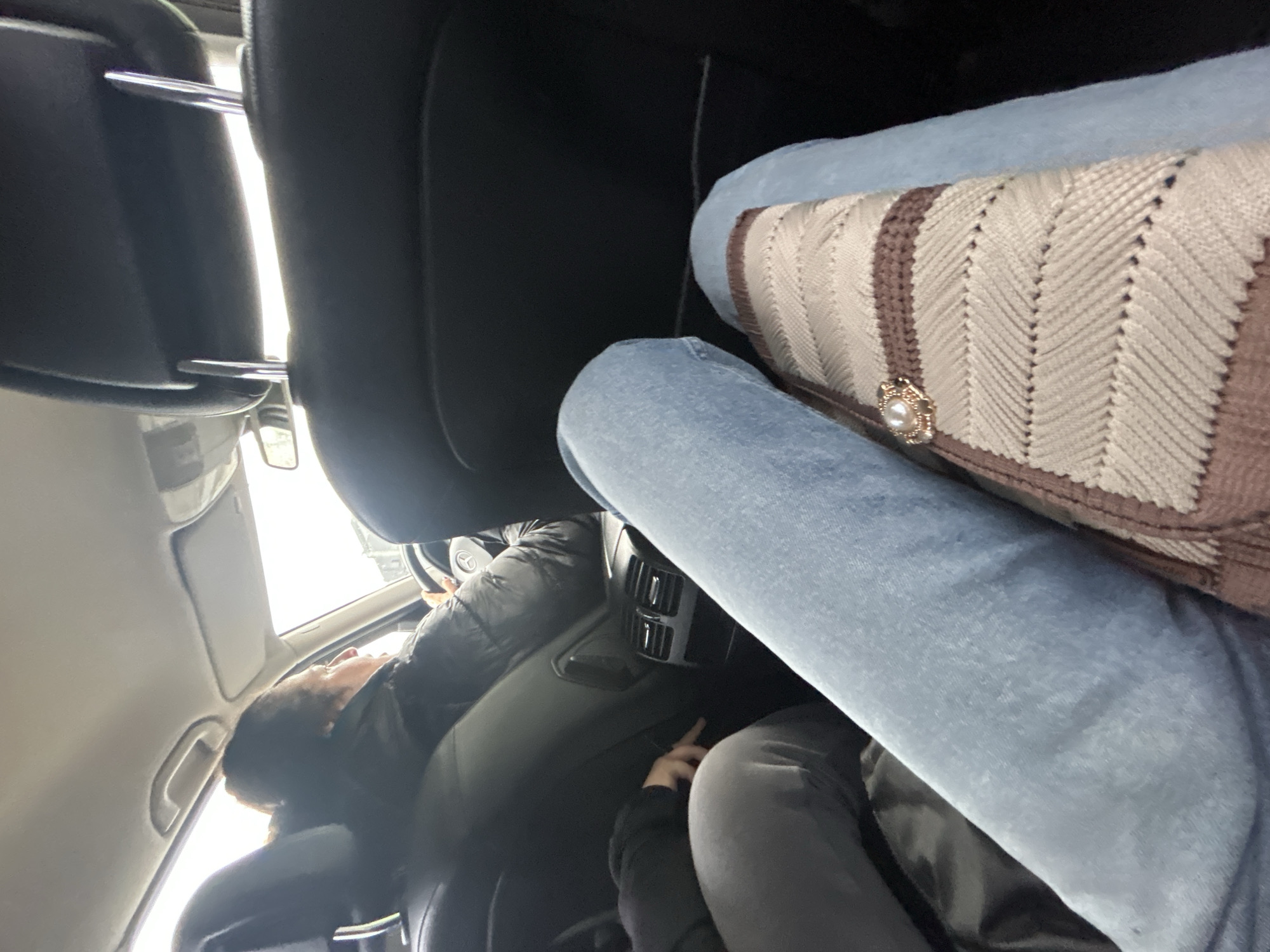 The interior of a vehicle is shown, with a person visible in the driver's seat. The driver is wearing a dark-colored jacket. A person's leg, clad in blue denim, is partially visible over the back seat. A patterned handbag, beige with brown accents and a decorative pearl-like ornament, sits atop the leg. The location in Ruse, Bulgaria is unknown.