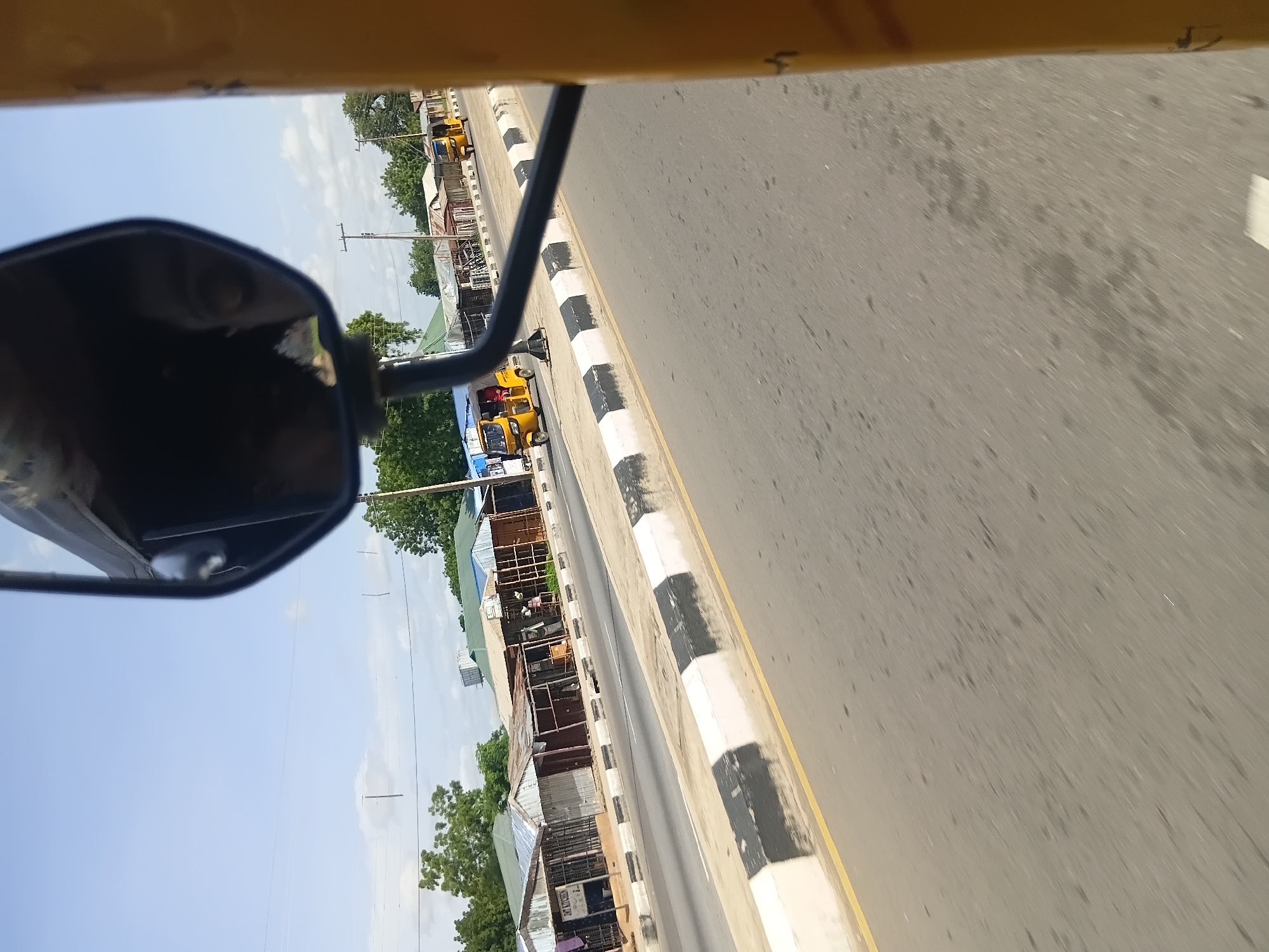 In Jalingo, Nigeria, on a bright day, a yellow tricycle moves along a paved road lined with small shops. The shops have corrugated metal roofs and walls, with some open to the street. A black and white striped barrier separates the road from the shops. The tricycle is in motion, and a person can be seen inside. The scene is viewed from another moving vehicle, with the side mirror of that vehicle visible in the foreground, reflecting the sky and the driver's face. The sky is partly cloudy, with a few wispy clouds against a clear blue. Trees are visible in the background, and utility poles dot the landscape.