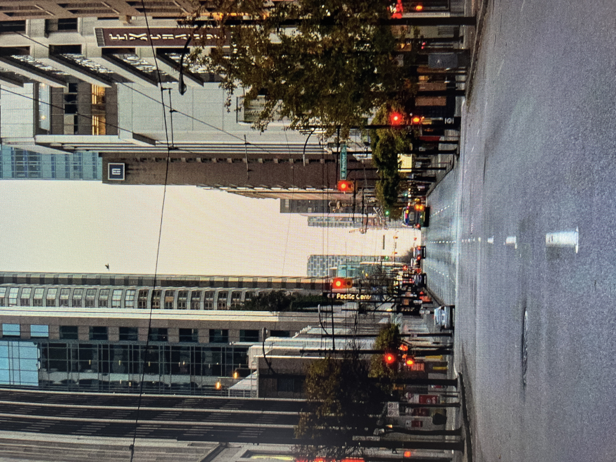 The image depicts an urban street scene, likely in Vancouver, Canada, based on the prominent "EXCHANGE" sign and street names visible. The perspective is looking down a wide, asphalt street lined with tall buildings on both sides. The buildings are a mix of modern glass-and-steel structures and older, more traditional architecture. Trees with autumn foliage line the sidewalks, adding a touch of color to the otherwise muted urban palette. Traffic lights are visible at intersections, with several showing red, indicating stopped traffic. A few cars are present on the road, as is a taxi. The sky is overcast, contributing to a soft, diffused light. The overall impression is one of a quiet city morning or late afternoon, with minimal pedestrian activity.