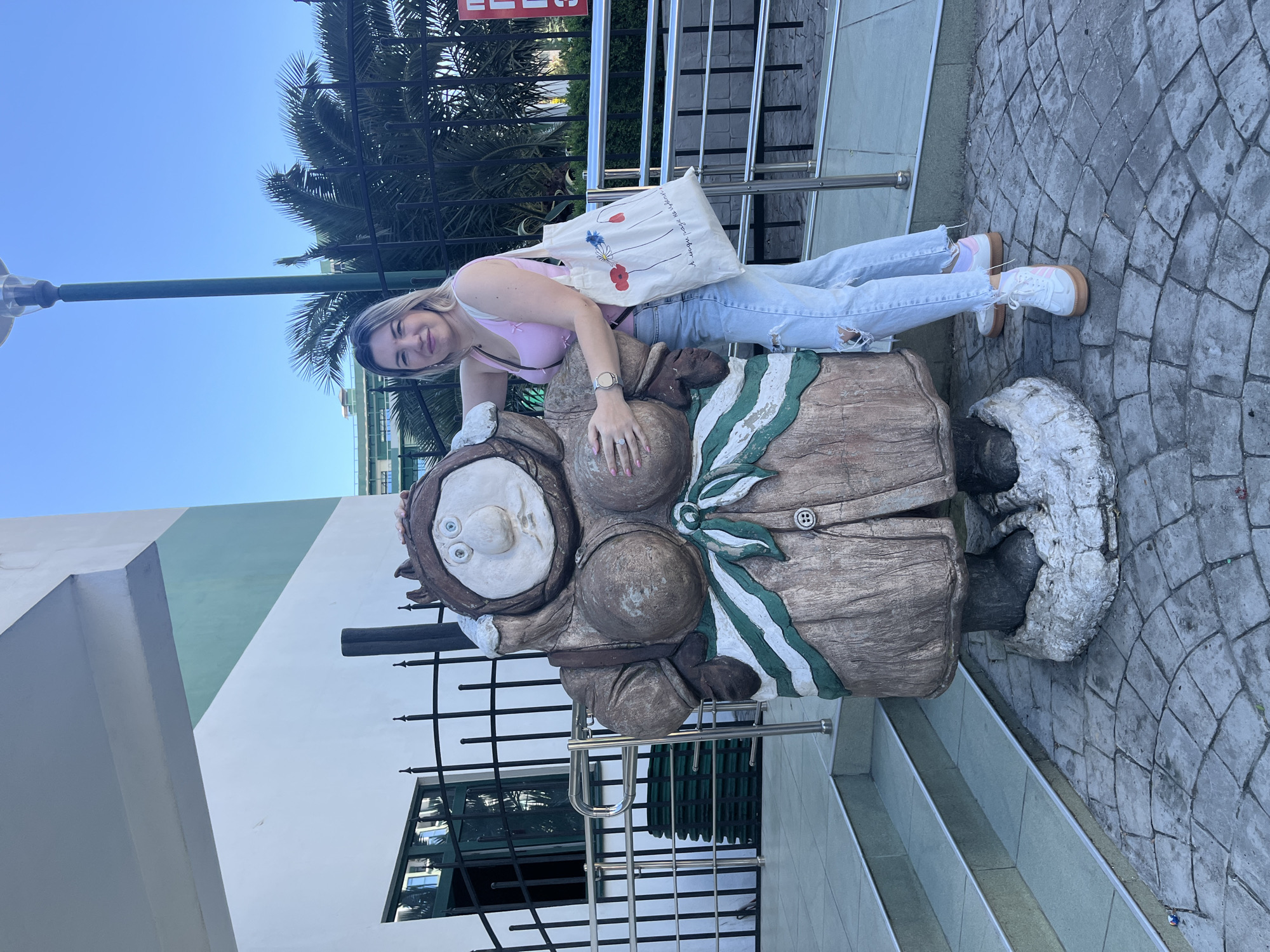 A young woman with blonde hair and bangs is posing for a photo next to a whimsical statue. She is wearing a light pink tank top and light blue ripped jeans, along with white sneakers. On her left shoulder, she carries a white tote bag adorned with a simple drawing of poppies. The statue, which appears to be made of stone or concrete, depicts a character with large, round eyes, a prominent nose, and a green and white striped scarf. The woman is leaning her head on the statue, with her left hand resting on its chest, and she is smiling at the camera.
The scene appears to be outdoors, with a clear blue sky visible in the background, suggesting a sunny day. Palm trees can be seen behind a black metal fence, hinting at a warm climate. The ground is paved with cobblestones, and there are steps leading up to the right of the image. A lamppost is also visible in the upper left corner. The woman's relaxed posture and smile convey a sense of happiness and enjoyment. While no specific time of day is explicitly indicated, the bright lighting suggests it is daytime, possibly mid-morning or afternoon. The location context of Sochi, Russia, aligns with the presence of palm trees and a sunny atmosphere.