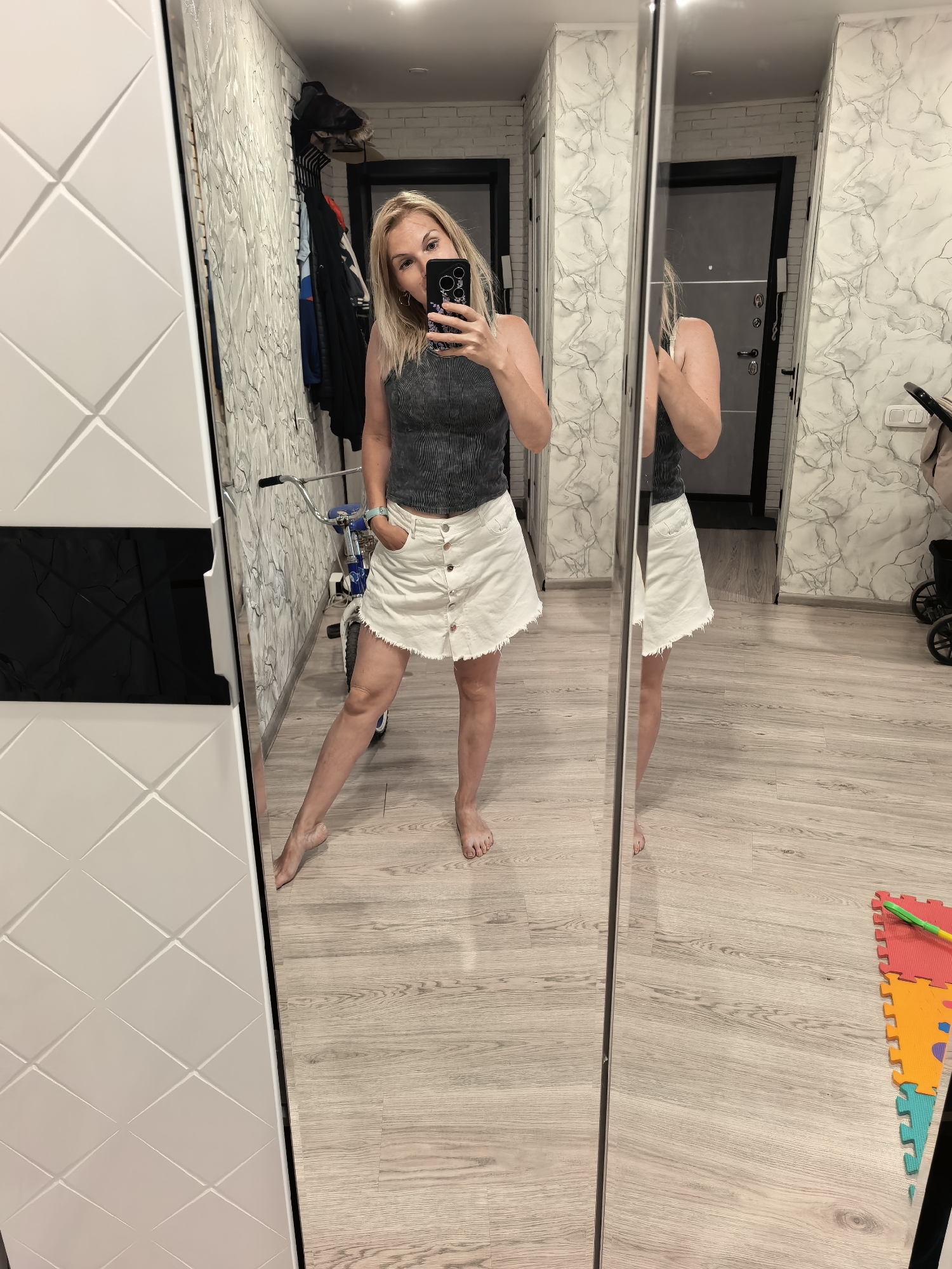 A woman is taking a selfie in front of a mirrored closet. She is wearing a gray tank top and a white skirt with buttons down the front. Her left hand is in her pocket. A child's bicycle is visible behind her. The interior decor features patterned wallpaper and wooden flooring. There are children's playmats on the floor. This scene is in Sterlitamak, Russia.