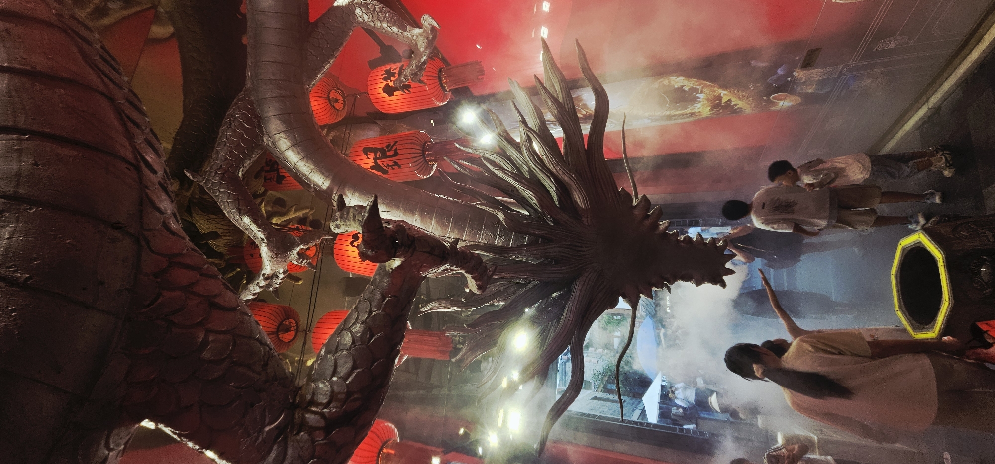 The image captures a dramatic indoor scene at the Yangtze River Cableway in Yuzhong District, China. Dominating the frame is a large, metallic dragon sculpture suspended from the ceiling, its head hanging down towards the viewer. Red, cylindrical lanterns with Chinese characters are visible in the background, emitting a warm glow that contrasts with the cooler tones of the dragon. The space is filled with a light fog or smoke, adding to the atmospheric effect.
In the foreground, several people are present, observing the spectacle. A woman with her hair pulled back in a ponytail stands facing the dragon, her arm extended as if pointing or interacting with the scene. To her right, two young boys are looking up at the dragon, seemingly captivated by its presence. Other figures are visible in the background, some holding cameras, suggesting they are tourists or attendees at an event.
The environment appears to be an exhibition space or a tourist attraction. The walls in the background show images and architectural elements, and there are signs and structures suggesting a well-developed public space. The overall impression is one of wonder and immersion in a cultural or festive display.