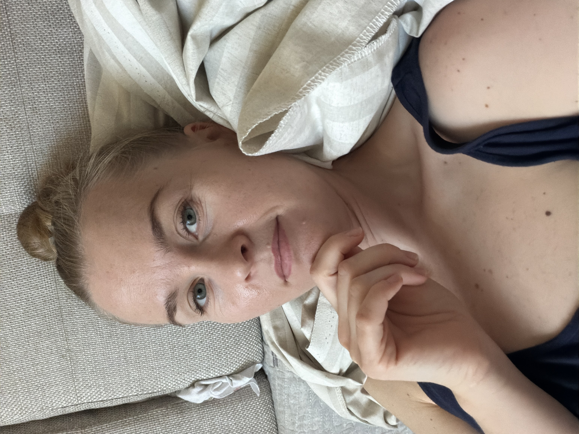 A woman is lying on a sofa, partially covered by a blanket. She is looking at the camera with her right hand touching her chin. Her hair is pulled up in a bun on top of her head. The sofa is upholstered in a textured, light-colored fabric. The blanket is white with subtle vertical stripes and has visible stitching along its edge. The overall lighting suggests an indoor setting. There is no identifiable indication of the city of Khabarovsk, Russia, within the image.