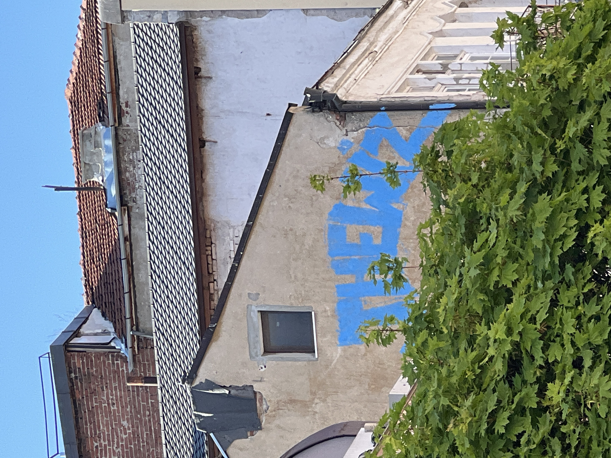 The image captures a building facade in an urban setting. The primary architectural elements include brick, stone, and stucco, with a mix of textures from tiled roofs to metal grating. A prominent blue graffiti tag is visible on the stucco surface of a gable end. A small, square window punctuates the gable wall. Decaying architectural details are apparent. A lush green tree fills the right side of the frame. The location is noted as Unknown, Unknown.