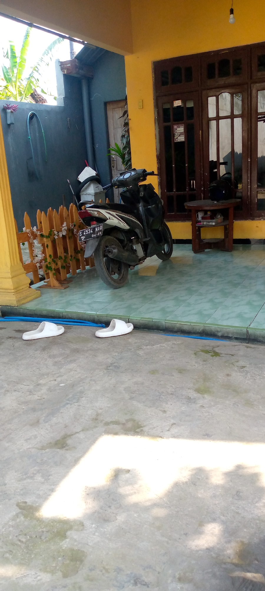 A still, eye-level, medium shot captures a residential porch area in Sumberwono, Indonesia. A black and white scooter is parked on the tiled floor, which is partially covered by a grey and blue tiled border. To the left of the scooter, a small, decorative wooden fence with white floral accents encloses a patch of greenery. Behind the fence, a dark grey wall with a blue hose and a white object is visible. A yellow building with brown-paneled doors and windows forms the backdrop on the right. The floor in the foreground is concrete and appears wet in patches, with shadows cast from an unseen light source. Two white flip-flops are placed on the concrete floor in the lower left portion of the frame.