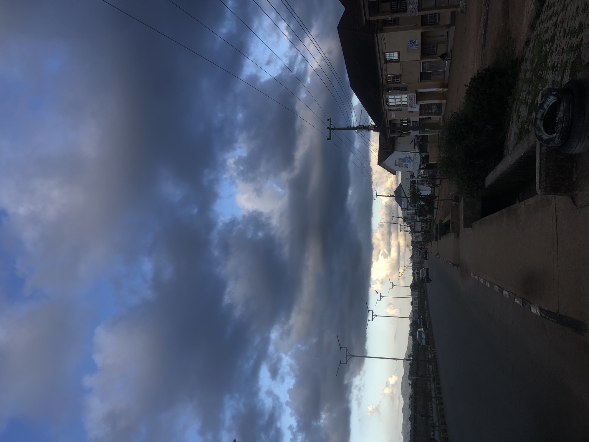 This image depicts a street scene in Barakin Lerawi, Nigeria, under a dramatic, cloudy sky. The sky is filled with large, dark, and grey clouds with patches of blue visible. The road, which appears to be paved, runs from the foreground left to the middle ground right, curving slightly. Several streetlights with a modern design are visible along the side of the road, casting minimal light due to the ambient conditions.
On the right side of the road, buildings with multiple stories line the street. These buildings have various architectural styles, some appearing residential and others commercial, with visible windows, doors, and balconies. One building has signage that reads "MINISTRY HOUSE." Some of these buildings are painted in light colors, while others have darker facades. Utility poles with multiple electrical wires crisscross the scene above the buildings and the road.
On the road, there are several vehicles, including cars and a motorcycle, in motion. Pedestrians are also present, walking on the sidewalk or near the road. The sidewalk on the right is made of concrete and appears to have a drainage channel running alongside it. In the foreground right, a pile of discarded tires is visible next to the drainage channel. In the background, a line of hills or mountains can be seen. The overall impression is of a street in a developing urban area, with a mix of infrastructure and daily life.