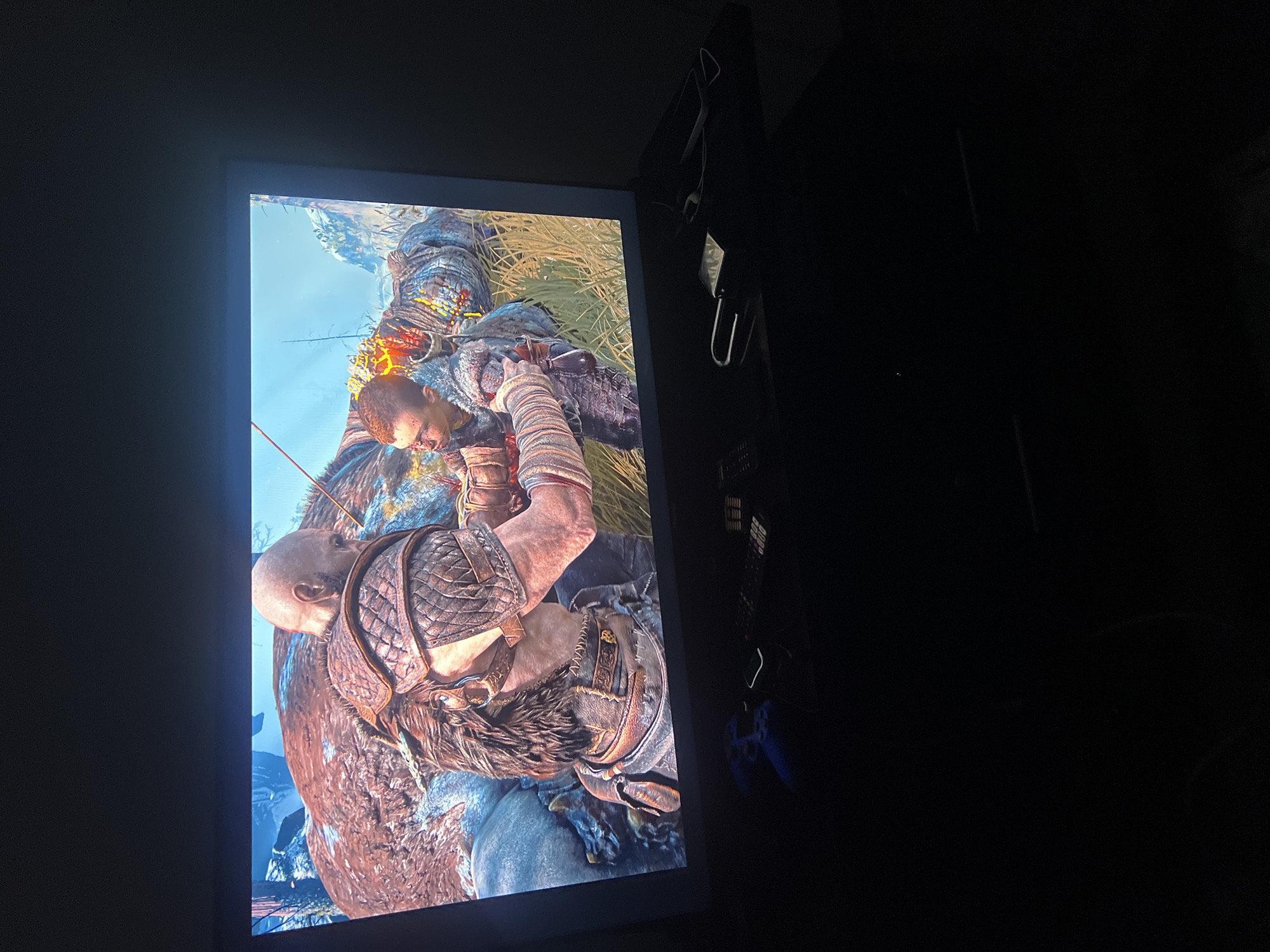 The image shows a television screen displaying a scene from a video game, likely God of War, featuring characters Kratos and Atreus. Kratos, a large muscular man with a bald head and beard, is wearing ornate armor and has his arm around Atreus, a younger boy in a fur-lined tunic. They appear to be interacting with a large, brown, furry creature that is lying on the ground. The background of the game scene depicts a grassy, mountainous landscape. Below the television, a gaming controller and a remote control are visible on a dark surface. The overall lighting suggests the image was taken in a dimly lit room. The image does not contain any information that would indicate its location in the city of Jos, Nigeria.