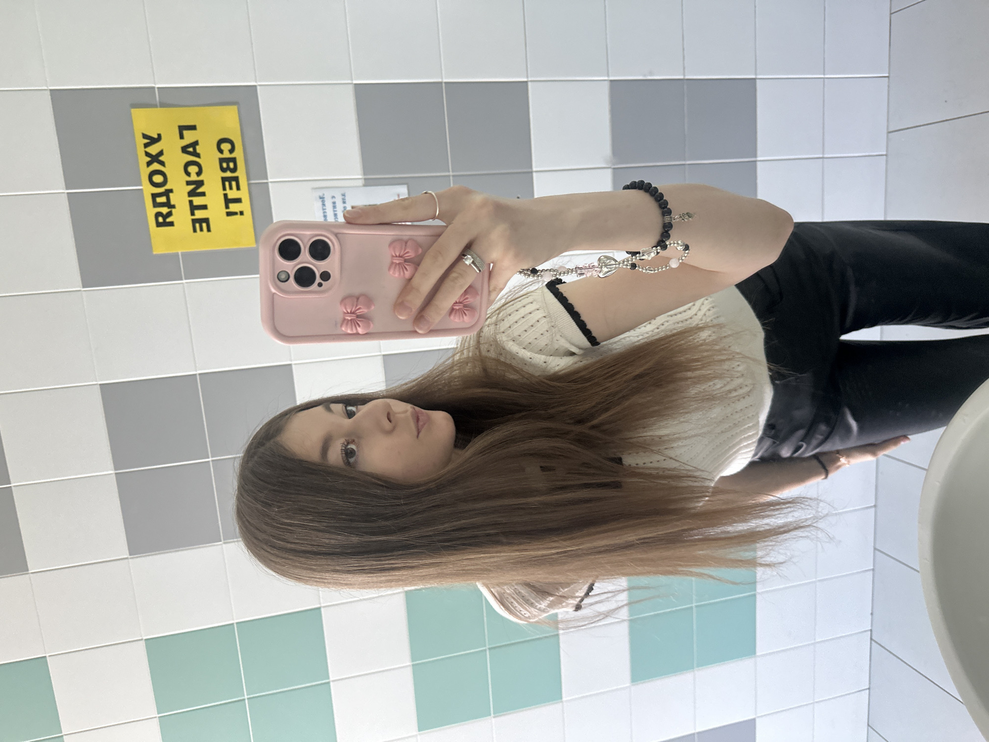 A young woman is taking a selfie in a bathroom, likely in Kazan, Russia, given the Cyrillic text ("СВЕТ! ЛУЧШЕ РЯДОМ!") on a yellow sign affixed to the tiled wall. She is holding a pink iPhone in her right hand, which has a decorative case with flower motifs. She has long brown hair. She is wearing a white top with black trim. She also has a bracelet and rings on her hands. The background consists of a tiled wall with alternating gray and white tiles, as well as some tiles with a greenish-blue tint. Part of a white sink is visible on the right side of the frame. She is wearing black pants.