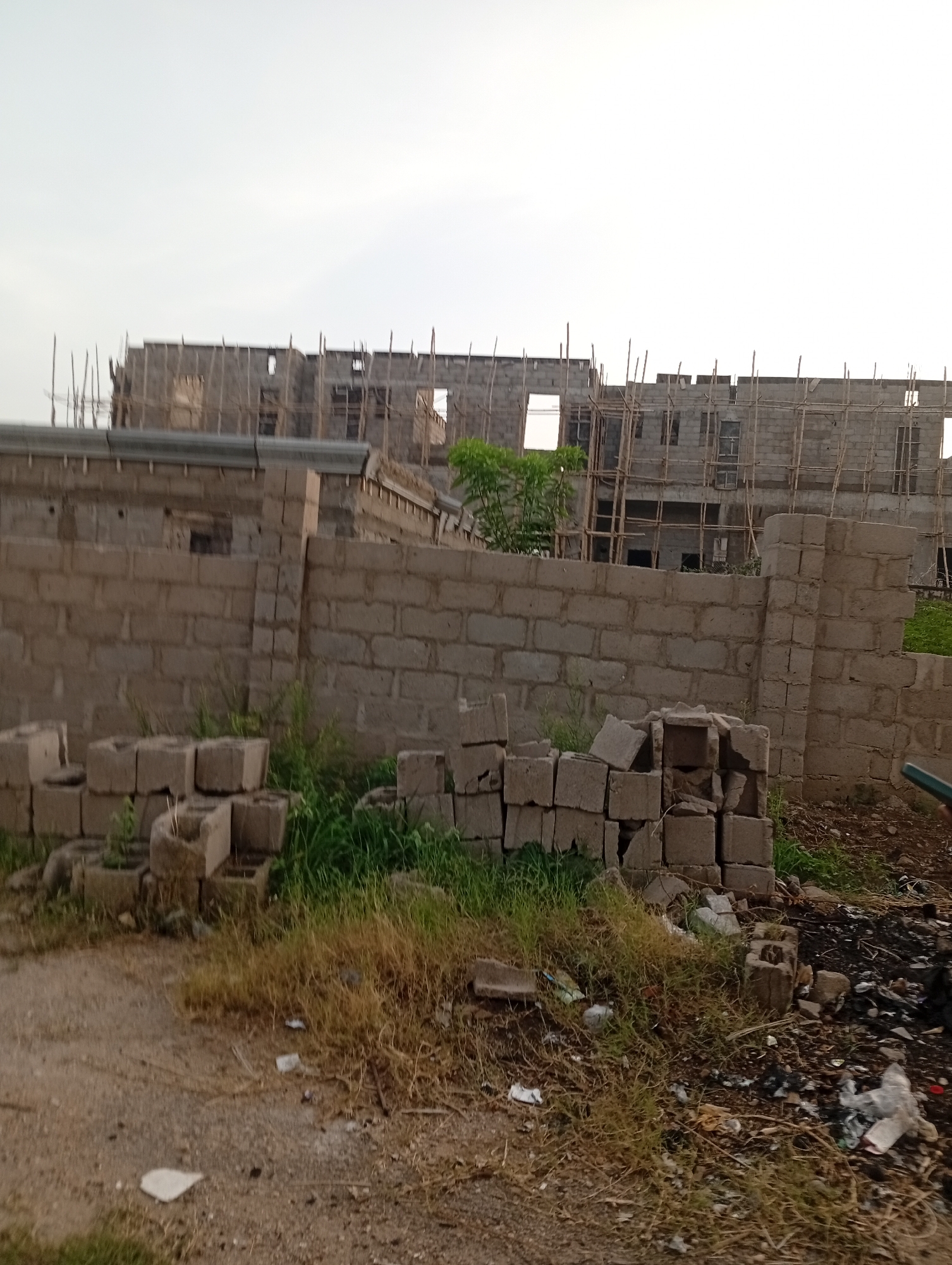 The image depicts a construction site in Adankolo, Nigeria. In the foreground, there are piles of gray concrete blocks scattered on the ground amid dry grass and patches of green vegetation. To the right, there's scattered trash, including plastic bags. A low concrete block wall stands partially constructed, acting as a boundary. Behind the wall, a larger building under construction is visible, supported by scaffolding made of wood poles. The sky is overcast, casting a muted light across the scene. No people are visible.