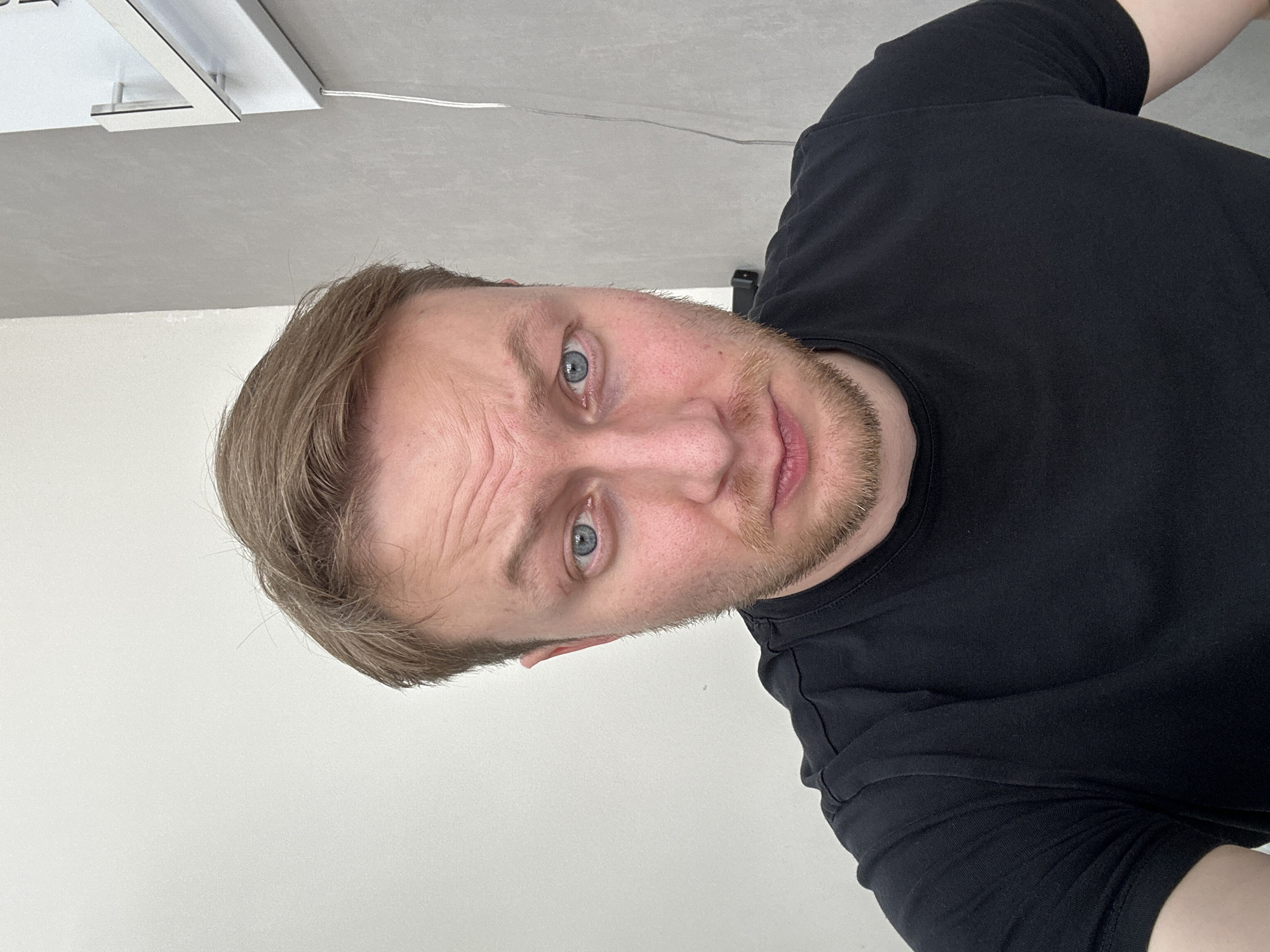 A man with short, light brown hair, blue eyes, and a stubbled beard is positioned in the center of the frame, wearing a black t-shirt. He is looking directly at the camera with a neutral expression. The background consists of a white wall and a ceiling with a white light fixture in the upper left corner. There are no other discernible subjects or objects present in the image, nor are there any visible elements to indicate the location is Sochi, Russia.