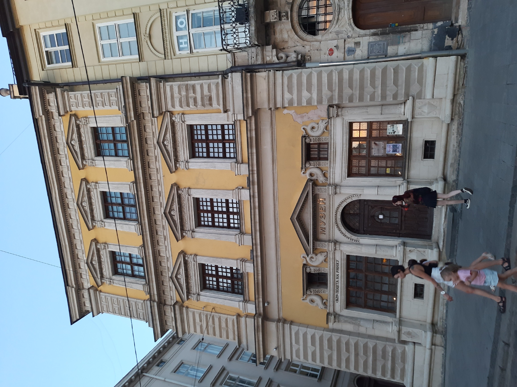 The image shows a street scene in Lviv, Ukraine. The main focus is the facade of a historic yellow building with ornate architectural details and multiple windows. Above an entrance, the word "МУЗЕЙ" (Museum) is visible in gold lettering. Below this, the text "•1629• ПОШТА НА РИНКУ" (•1629• Post Office on the Market Square) is inscribed. To the right of the yellow building, a neighboring building with a balcony and windows is visible. In the foreground, three people are walking on the sidewalk. A woman in a black top and white skirt walks with a child in a pink top and blue skirt. Another woman with long red hair walks behind them. The sky is clear and appears to be dusk. The street is paved.