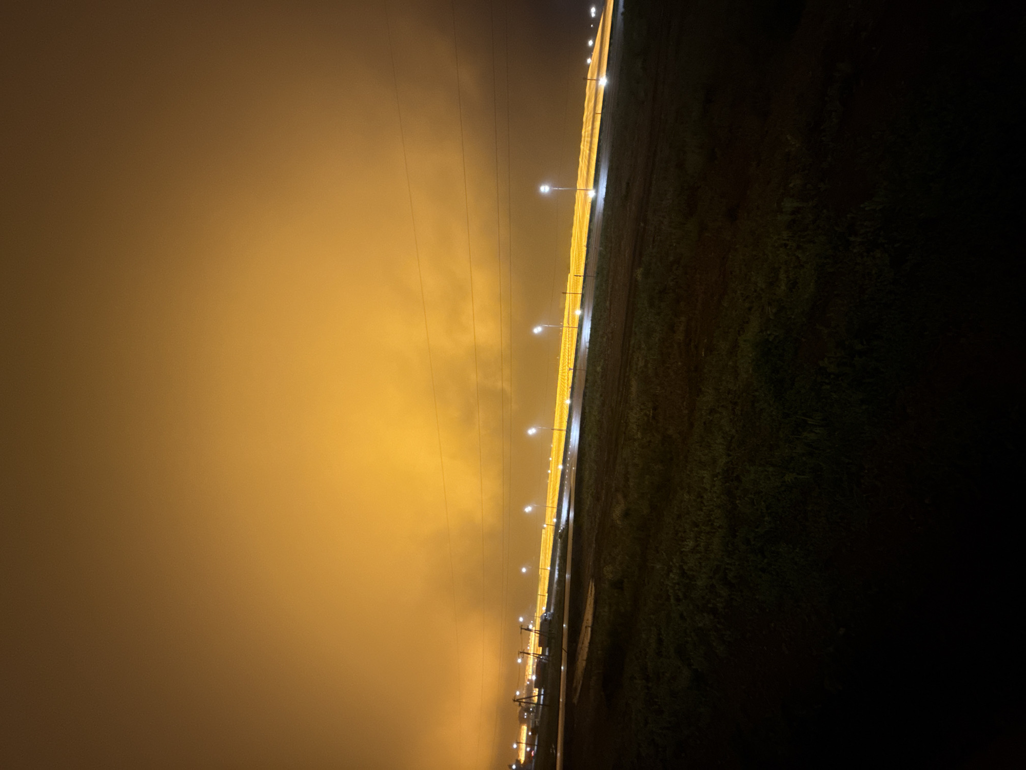 A low-angle nighttime photograph captures an illuminated roadway in Sarapul, Russia, under an intensely glowing orange-yellow sky. The upper two-thirds of the frame are dominated by a dense, hazy orange-yellow cloud cover, reflecting significant light pollution from below. Horizontally bisecting the midground is a long, brightly illuminated stretch of roadway infrastructure, characterized by a continuous, intense yellow luminescence. Numerous white-light street lamps are distinctly visible along its length, contributing to the overall illumination. Above the roadway, multiple dark overhead utility lines traverse the frame, supported by utility poles partially visible on the left. The foreground presents a dark, uncultivated terrain with sparse, low-lying vegetation. Portions of the ground appear damp or puddled, reflecting minimal ambient light. No human figures, animals, or active vehicles are explicitly discernible.