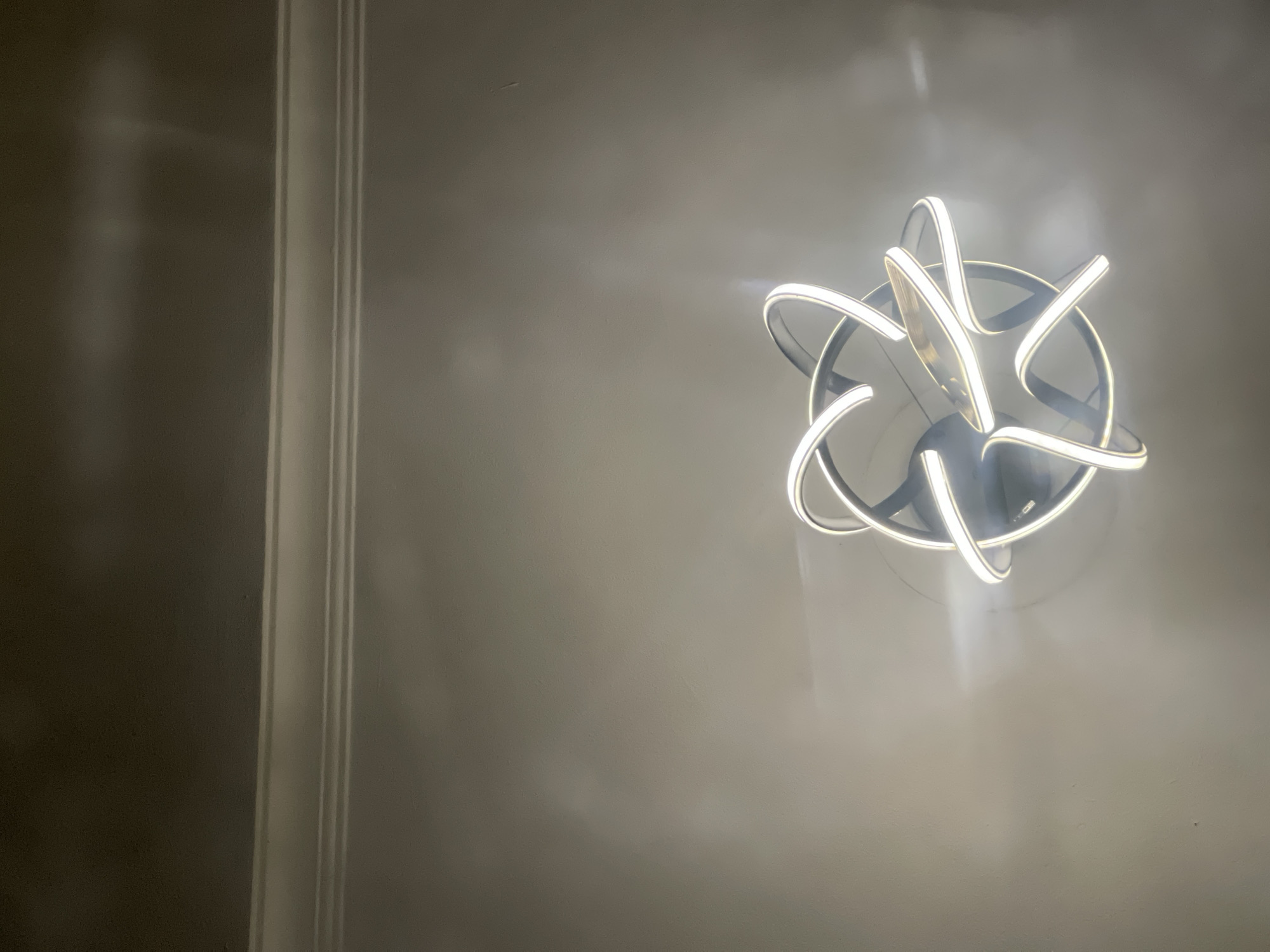 The image shows an interior view of a room. A modern ceiling light fixture, featuring circular and curved light elements, is positioned against a light gray or off-white ceiling. The lighting fixture emits a bright, warm light. The left side of the image shows a portion of a wall with a vertical architectural molding detail. The wall is painted or finished in a similar light gray or off-white tone as the ceiling. There are no people or interactions visible in the image. The location is not discernible from the given image and cannot be pinpointed to Abuja, Nigeria.