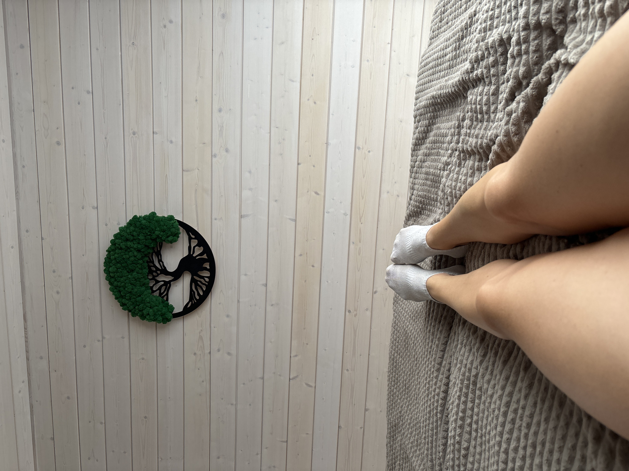The image shows a person's legs and feet, wearing white socks, resting on a textured gray blanket. The legs are positioned on the right side of the frame, with the feet pointing towards the bottom. The background on the left is a white wooden plank wall. On the wall, to the left of the legs, is a circular wall decoration. The decoration features a black silhouette of a tree with intricate branches within a circle, and one half of the tree is covered in vibrant green moss. The lighting appears to be natural and soft, suggesting it could be daytime. There is no visible text in the image. The scene appears to be indoors, possibly a bedroom, given the presence of the blanket and wall decor. The overall mood is relaxed and peaceful.