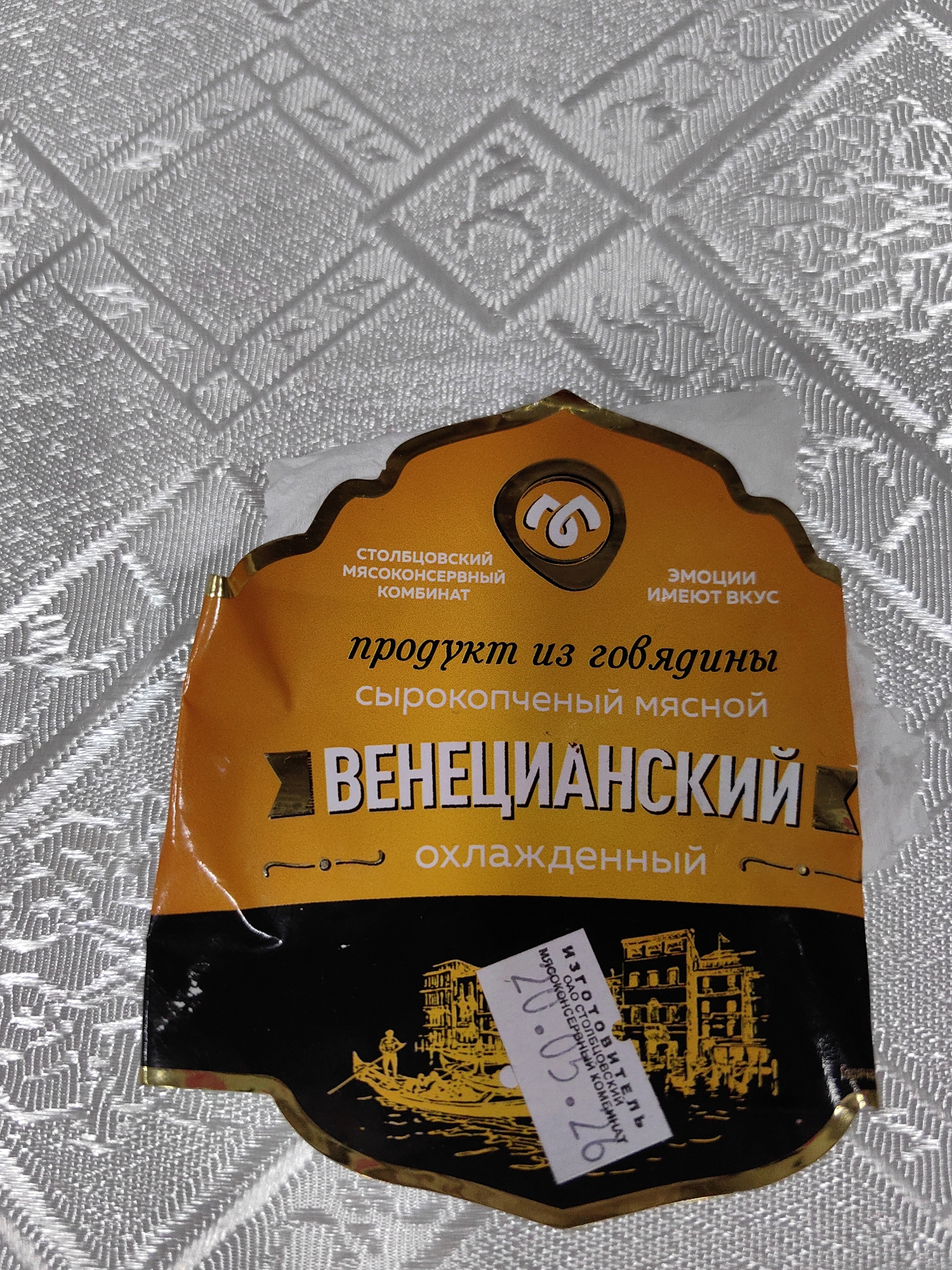 The image shows a close-up of a food product label with Russian text, set against a white, textured background. The label is predominantly orange and black, featuring a stylized logo, text describing the product as "product from beef, smoked meat," and the name "VENETSIANSKY." Below the main text, it says "chilled." There is a small vignette of a gondola and buildings on the black portion of the label. A white sticker with Russian text and a date "20.05.26" is affixed to the lower right corner of the label. The product is not located in any specific city.