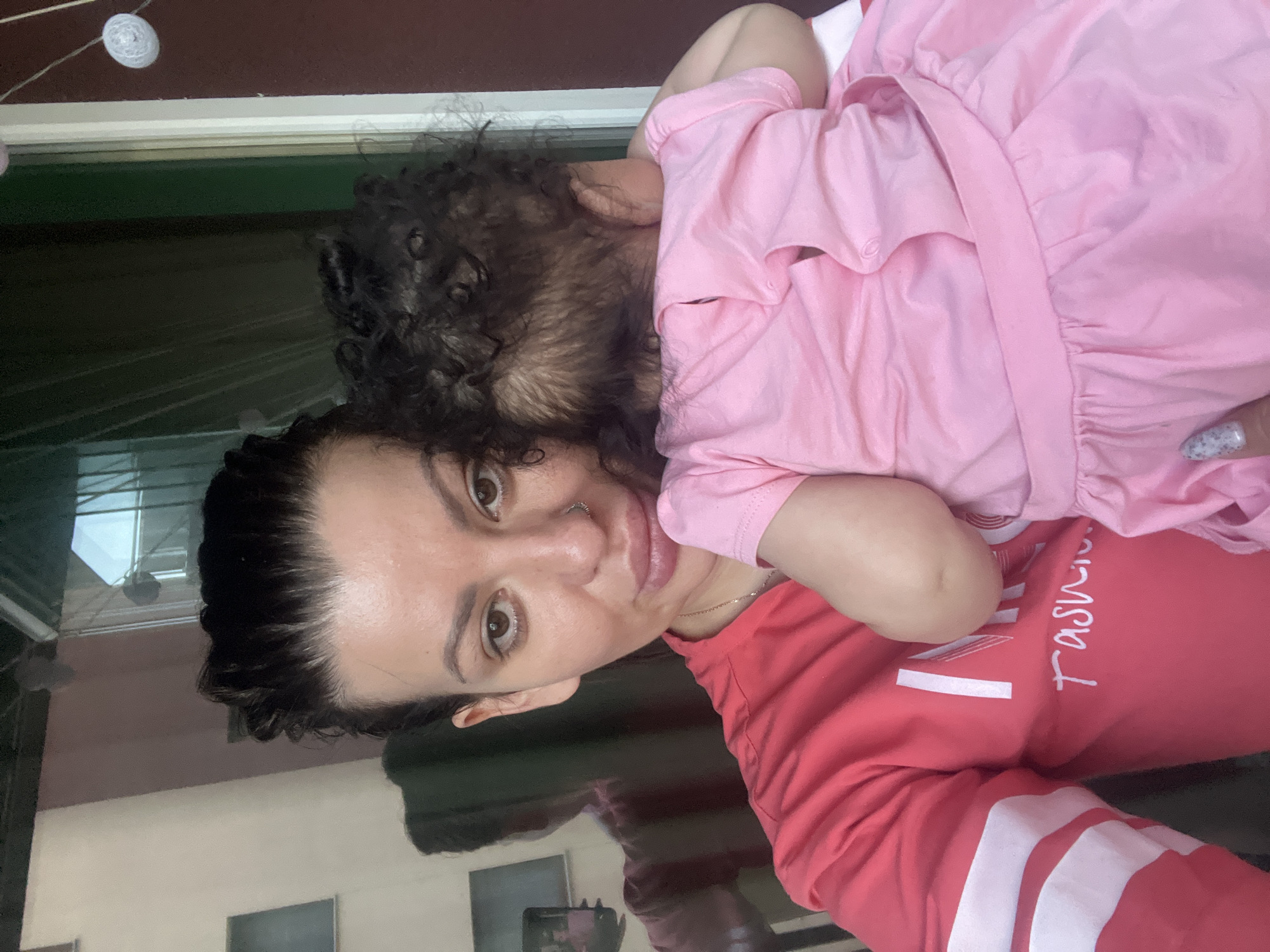 A woman with dark hair and a nose ring is holding a baby. The woman is wearing a pink sweatshirt with white stripes on the sleeves and white lettering that appears to say "Fashion". The baby is wearing a pink dress. The background appears to be an outdoor patio with glass doors and a railing. There is a reflection of the woman and baby in the glass. The image was taken on a balcony in Plovdiv, Bulgaria.