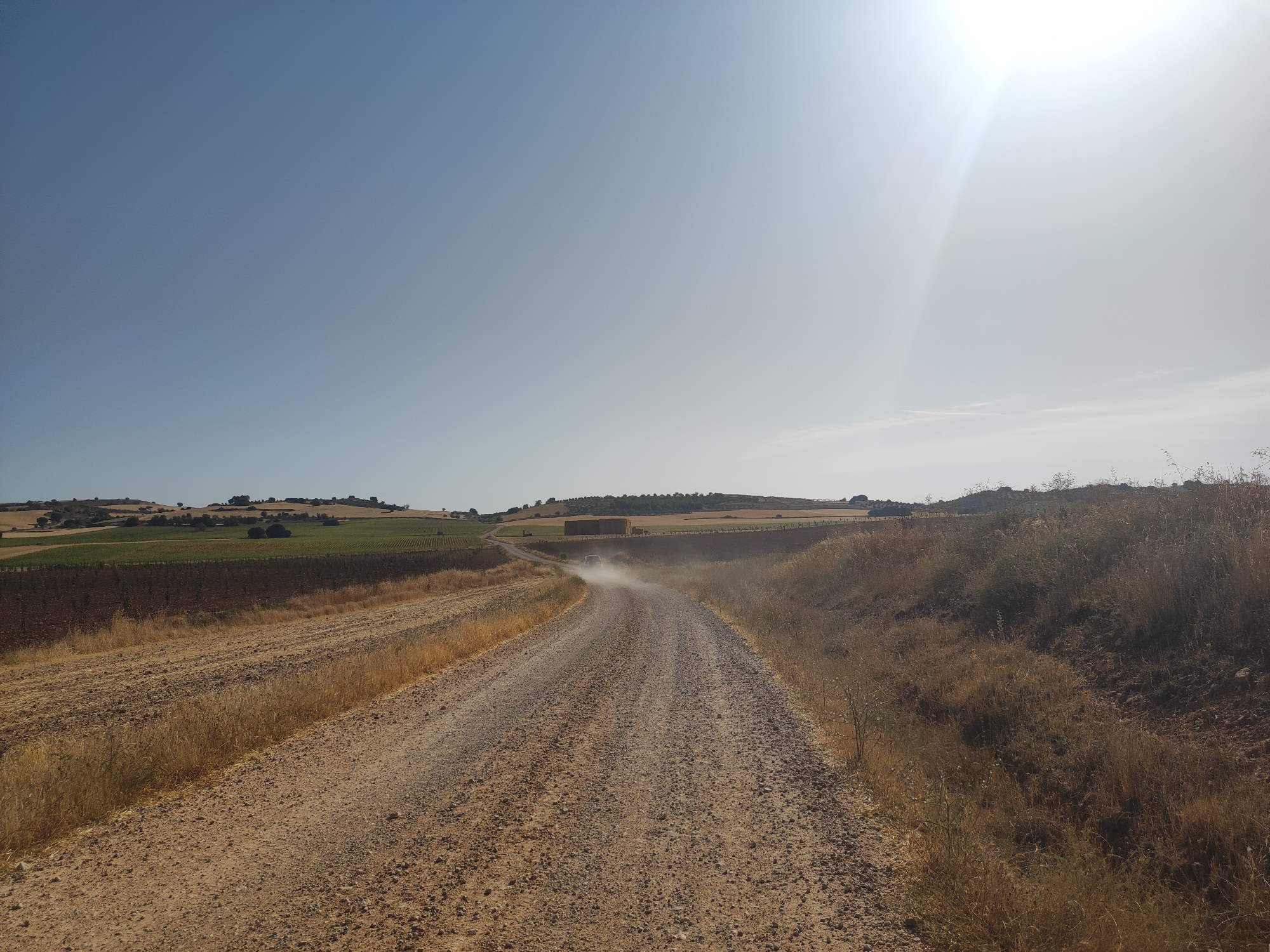 This is a wide-angle, eye-level, outdoor, daytime photograph of a dusty, unpaved road curving through a rural landscape under a clear, bright sky. The sun is high and to the right, casting a strong glare and lens flare into the upper right quadrant of the image. The road is made of dirt and gravel and is heavily eroded in places, with visible tire tracks. A small, dark-colored vehicle is visible in the distance on the road, kicking up a significant cloud of dust as it travels away from the camera.
The surrounding landscape consists of rolling hills and fields. To the left of the road, there are fields of dry, golden grass and stubble, suggesting a harvested crop. Further in the distance on the left, there are patches of green fields, possibly vineyards or crops, and some scattered trees. To the right of the road, the terrain is rougher and covered in dry, sparse vegetation and weeds. In the far distance, on the horizon, are more rolling hills dotted with trees and some scattered structures. Several large, rectangular hay bales are visible in the mid-ground on the left, beyond the green fields.
The scene appears to be in an agricultural area, likely during a dry season given the dust and the appearance of the fields. The specific location is identified as Villarejo de Fuentes, Spain. The overall impression is one of a remote, sun-baked rural environment.