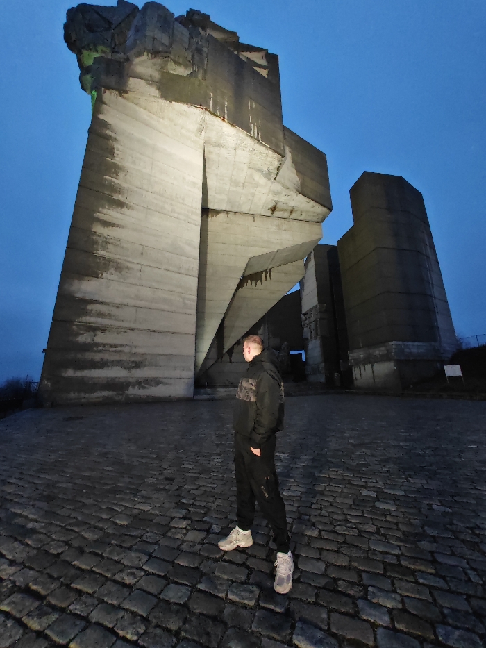 The image displays a young adult male standing on a dark cobblestone plaza in front of the Monument to 1300 Years of Bulgaria, located in Shumen, Bulgaria. The scene is illuminated by the ambient light of twilight or early night, indicated by a clear, dark blue sky.
The male individual is positioned slightly right of the frame's center, facing generally towards the right with his head turned to observe the monument. He is dressed in a black hooded jacket featuring a camouflage pattern across the chest and upper arms, dark (likely black) cargo-style trousers, and light-colored athletic shoes. His hands are not clearly visible, possibly in pockets.
The monument is a large, multi-component Brutalist concrete structure dominating the background. Its central, most prominent section consists of towering, angular concrete slabs, illuminated from below by artificial light, revealing vertical streaks and surface discoloration. The uppermost part of this central element exhibits a distinct green illumination. To the right and slightly behind this central structure, several darker, more cylindrical or block-like concrete forms are visible. The monument's surfaces appear weathered.
The ground beneath the individual and extending towards the monument is paved with irregular dark cobblestones. A small, light-colored rectangular sign is partially visible on the ground plane to the far right of the monument's base.
