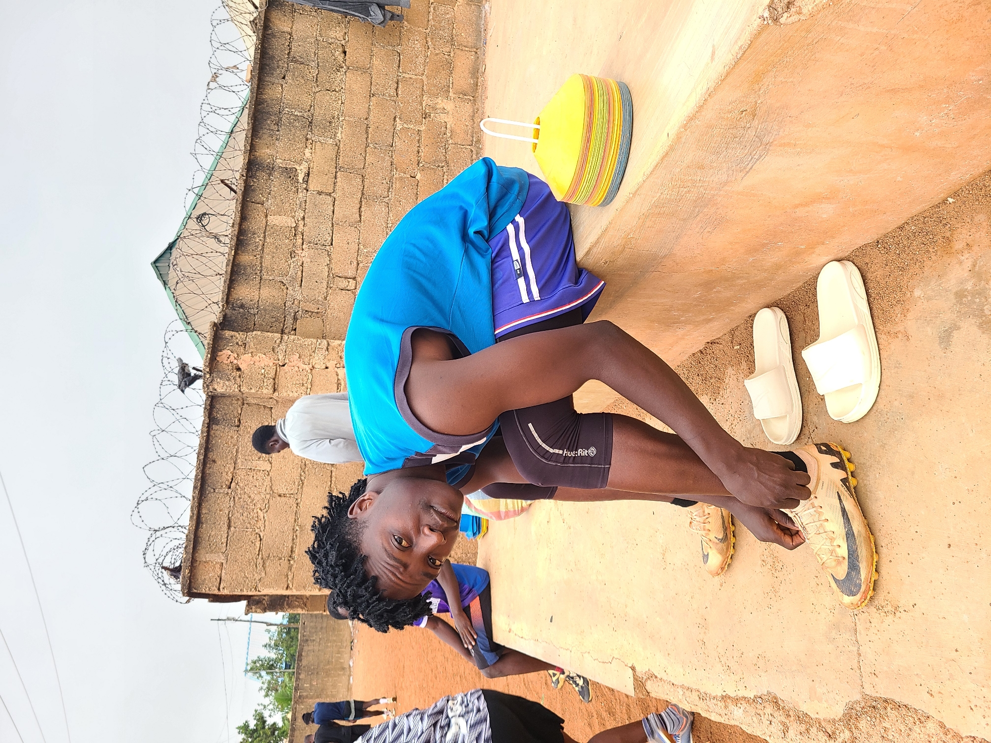 A young man is crouched down tying the laces of his yellow and black soccer cleats on a concrete surface in Barakin Lerawi, Nigeria. To his left is a stack of yellow and blue cones. Behind him are two pairs of white sandals. In the background are other people, a brick wall topped with barbed wire, and trees.