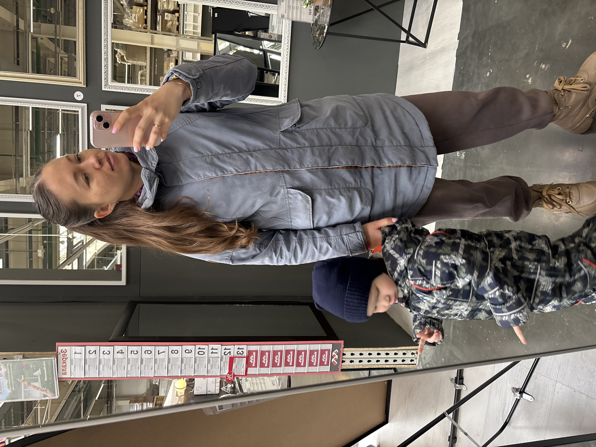 The image captures a woman taking a selfie in a store, possibly a home goods store, with her young child standing beside her. The location is likely Perm, Russia, based on the signage with Cyrillic script visible on a promotional display. The woman is holding a pink smartphone to take the picture. She is wearing a gray jacket, brown pants, and tan boots. Her long brown hair is visible. The child is wearing a navy blue beanie and a patterned jacket. They are standing next to the woman. The reflections in the mirror add to the complexity of the image, showing more of the store's interior. In the background, there are shelves with mirrors. A sales tag is visible, bearing price numbers, likely indicating products on sale. The floor is a neutral gray color.
