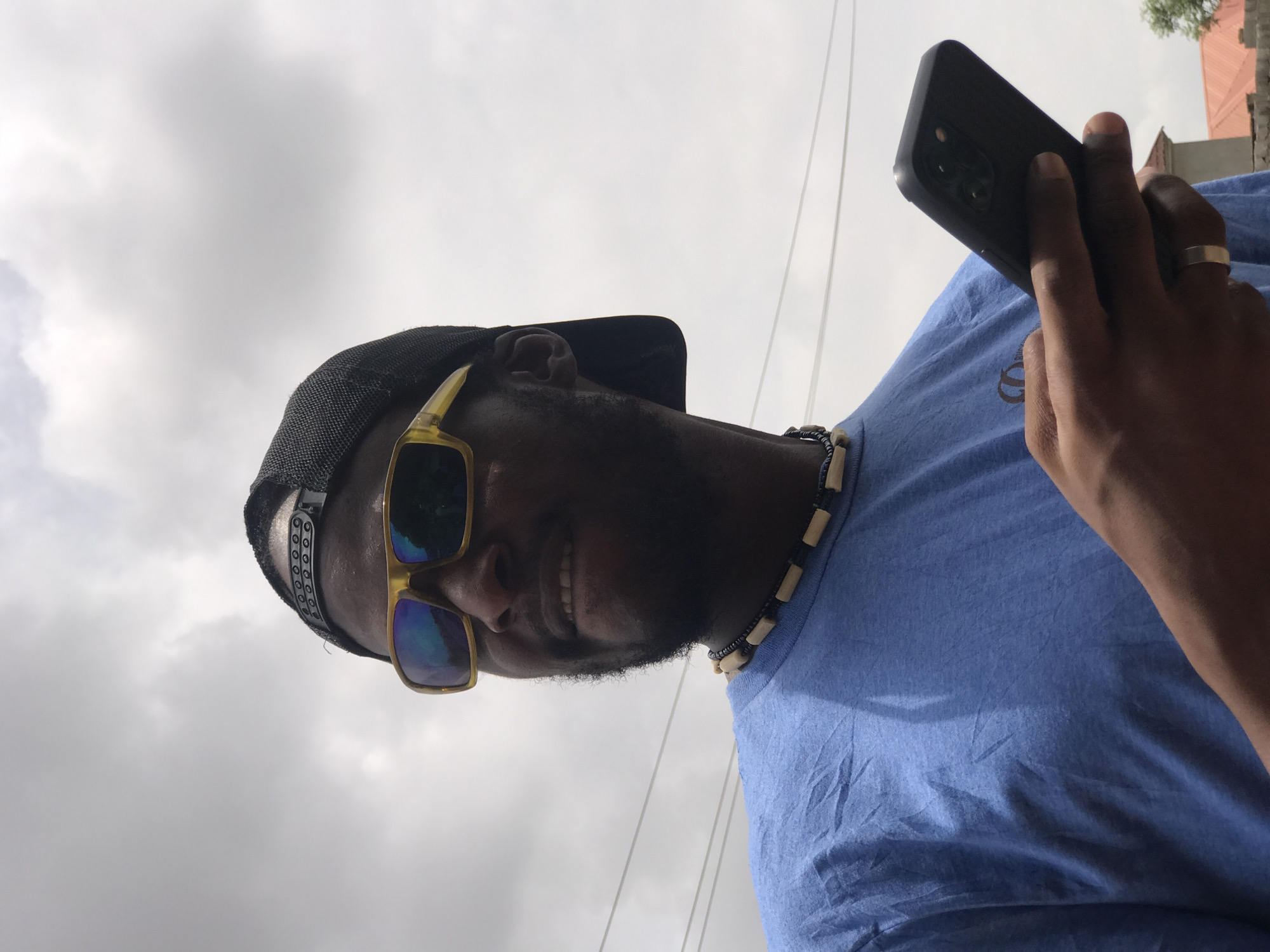A dark-skinned man with a beard and wearing a black cap, gold sunglasses, and a blue t-shirt is holding a black smartphone. He is also wearing a beaded necklace and a ring on his left hand. The background shows a cloudy sky and the tops of buildings. The location is implied to be in Abuja, Nigeria.