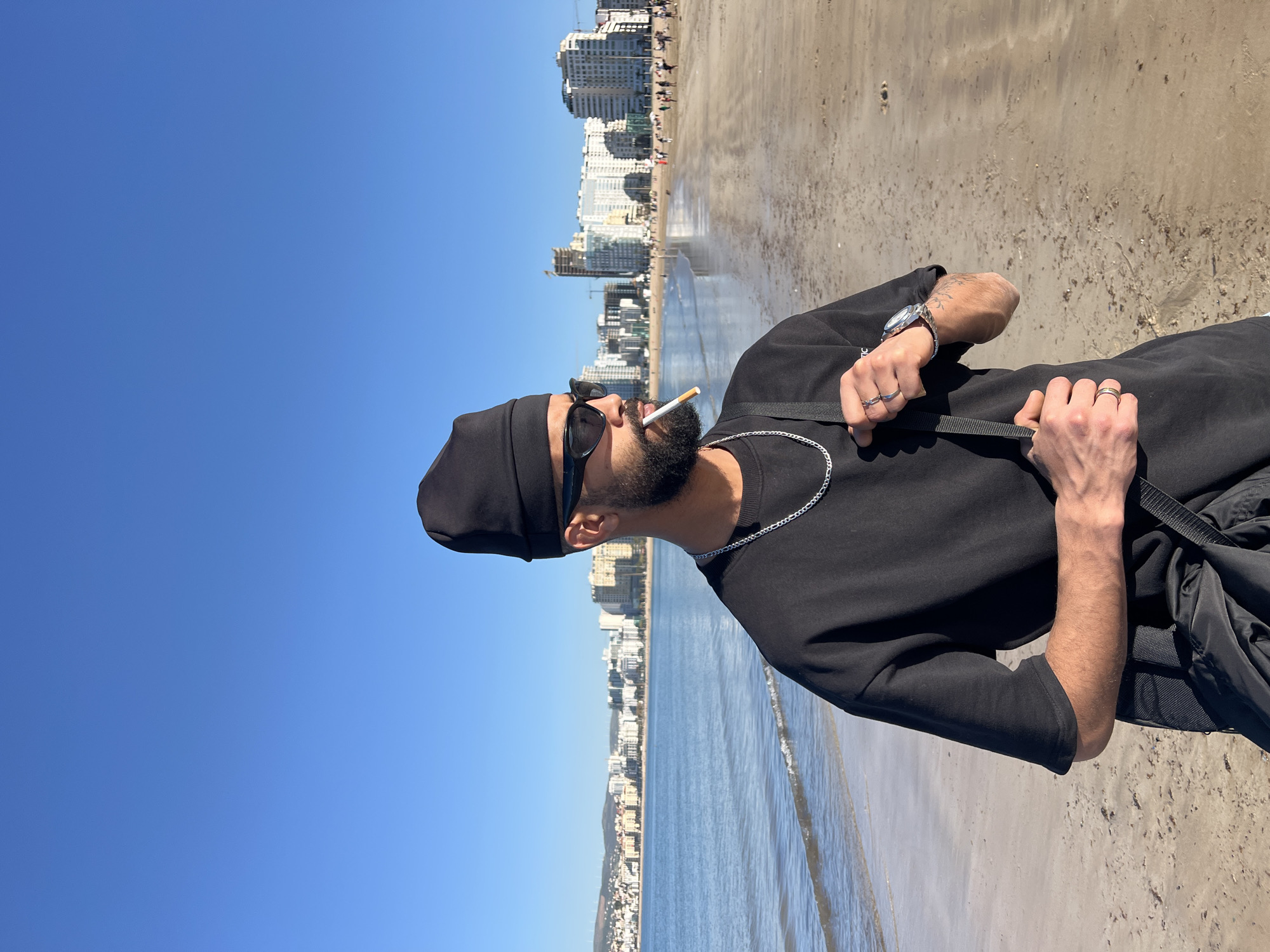 A man wearing a black hat, black sunglasses, and a black sweatshirt is standing on a beach with a cigarette in his mouth. He is facing left, with his back to the ocean. The city of Tanger طنجة, Morocco, is visible in the background. The sky is blue and the sand is tan. The man is holding a black bag with a strap. He is also wearing a silver watch and several rings.