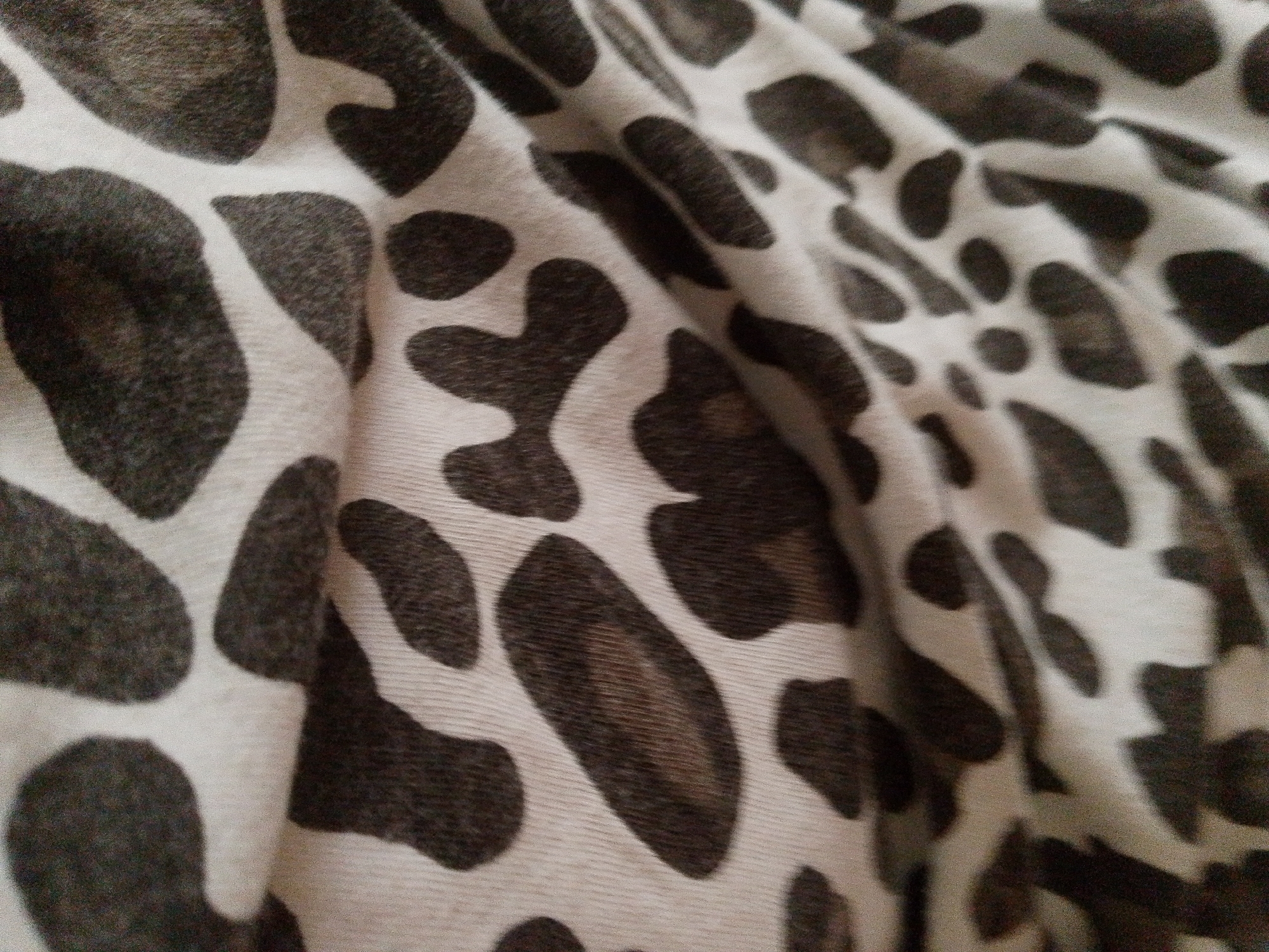 This is a close-up, slightly blurry photograph of a white fabric with a dark brown or black pattern. The pattern consists of irregular, blotchy shapes that resemble animal spots or a dalmatian print. The fabric is creased and folded, creating shadows and highlights that give it a three-dimensional appearance. The texture of the fabric appears to be soft, possibly like a knit or a faux fur, due to the fuzzy edges of the dark spots. The lighting is dim, and the overall impression is one of a textured textile surface. There is no identifiable scene, setting, people, or any visible text in the image. The image appears to be taken indoors, with no indication of the time of day or weather.