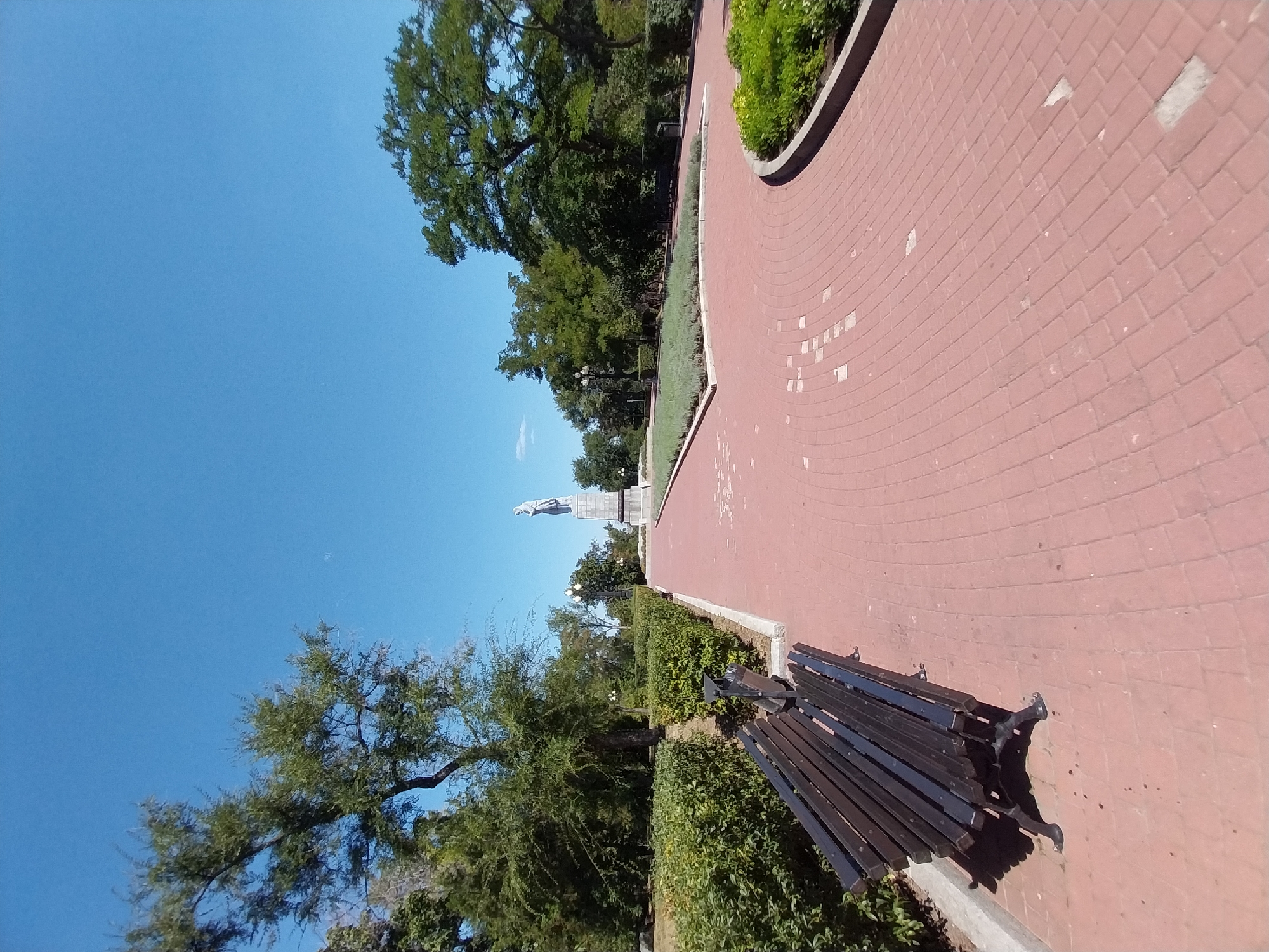 The image depicts a park pathway with a monument in the distance. In the foreground, a black wooden bench with metal legs is positioned on a red brick walkway. To the left of the bench, manicured green hedges form a border. The walkway curves to the right, leading towards a monument consisting of a statue atop a pedestal, situated in a green area with lampposts. Dense trees with green foliage surround the monument and the pathway, under a clear blue sky. The scene appears to be in the city of Dnipro, Ukraine, specifically at the Monument of Glory on Glory Square.