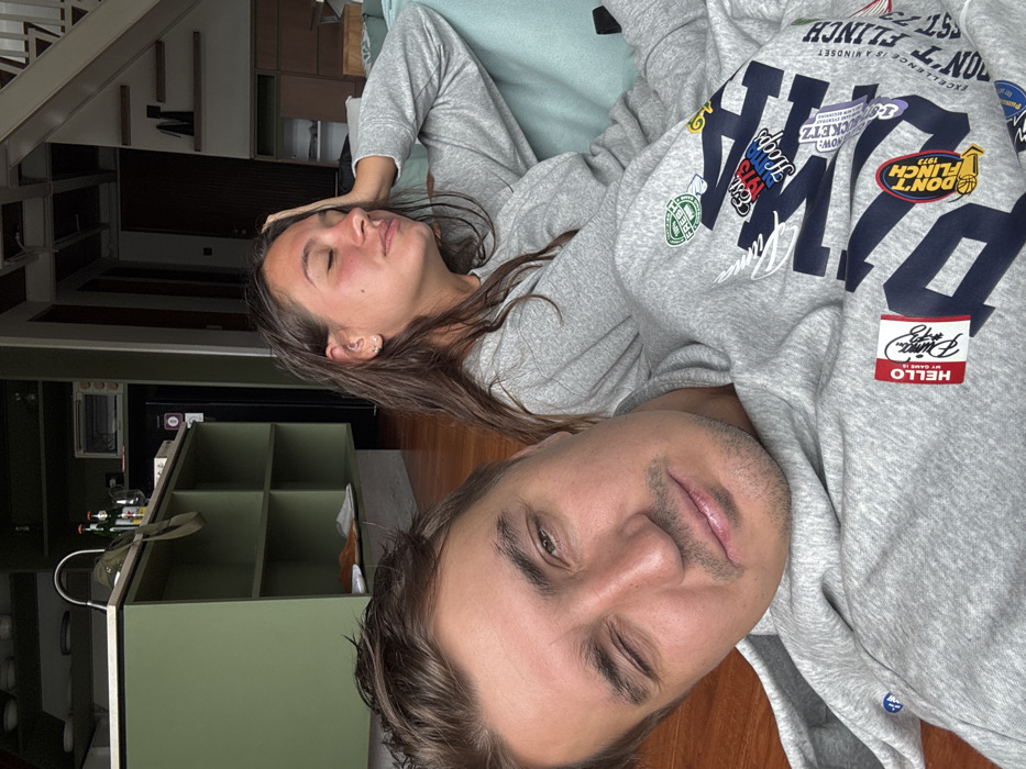 A couple, possibly on a couch, is captured in a rotated selfie. The woman, with closed eyes, rests her head. The man winks at the camera, revealing light stubble and neatly styled hair. Both are dressed in matching grey sweatshirts. The man's sweatshirt has a prominent "Puma" logo adorned with various patches and phrases such as "Don't Flinch" and "Hello My Name Is." Behind them, part of an organized home is visible with shelves, bottles, and a TV. The overall image is relaxed and intimate.