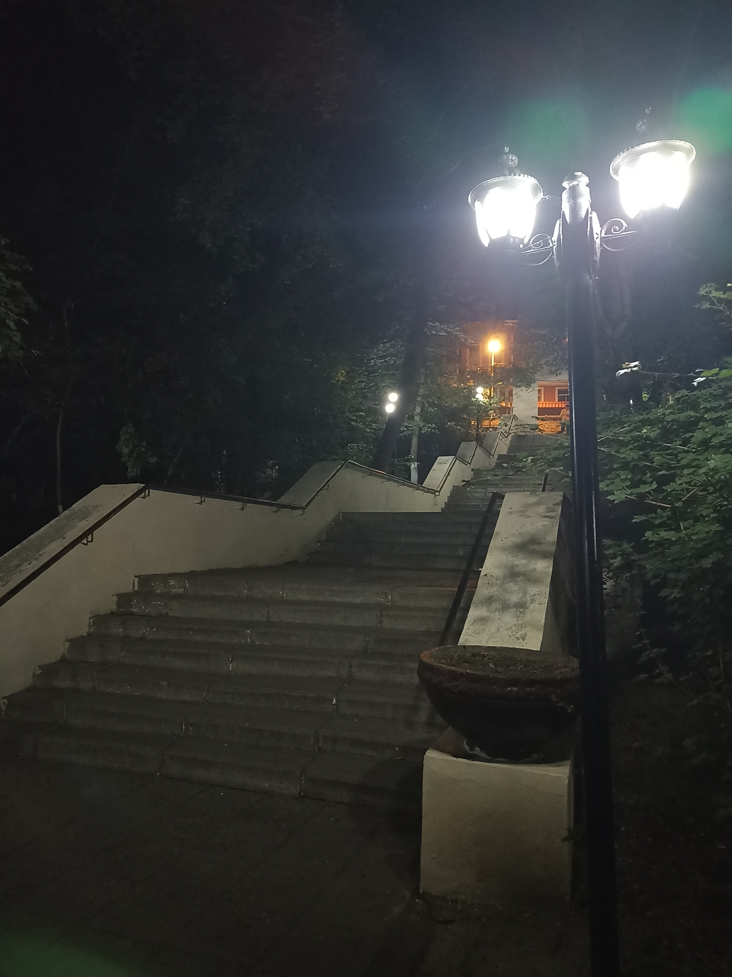 The image depicts a nighttime view of a grand stone staircase with a concrete balustrade and handrails, ascending into darkness. A vintage street lamp with multiple globes illuminates the scene, casting a warm glow on the steps and surrounding foliage. In the background, another light source, possibly from a building, can be seen further up the stairs. The stairs are flanked by dense trees and bushes, creating a natural and somewhat secluded atmosphere. The overall impression is one of quiet, late-night ambiance in a park or public space. The location is identified as Gusar, Azerbaijan.