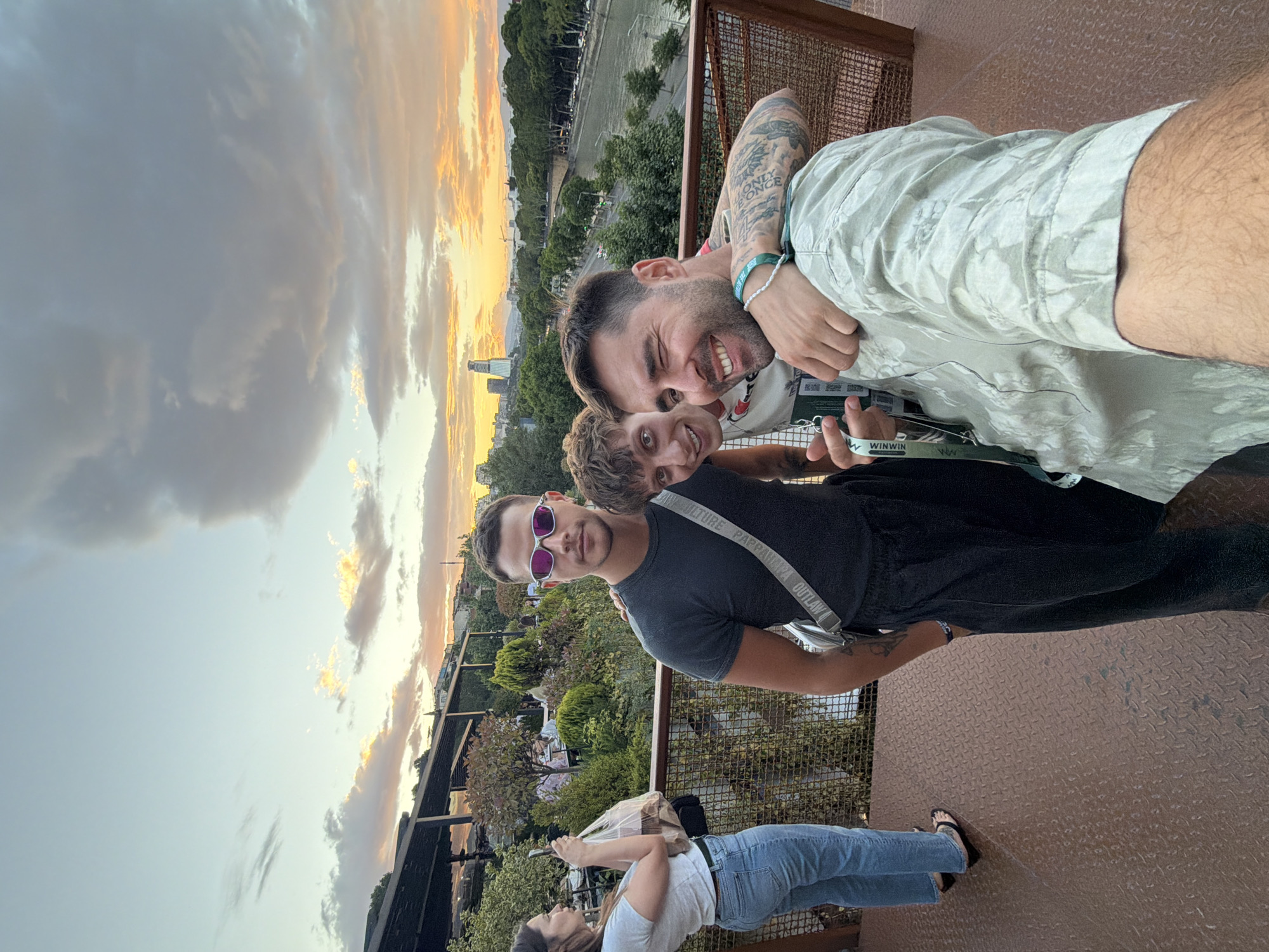 In Tbilisi, Georgia, at sunset, three young men pose for a selfie on a metal platform overlooking a city skyline. The man in the foreground, on the right, winks at the camera, his arm around another man's shoulder. He wears a light green patterned shirt and has tattoos on his arm. The man in the middle, with curly hair, smiles broadly. The man on the left, wearing pink-tinted sunglasses and a dark t-shirt, stands stoically, a silver crossbody bag slung over his shoulder. In the background, a woman in a white t-shirt and jeans holds a phone, seemingly taking a picture of the group or the scenery. The sky is dramatic with orange and grey clouds. Trees and buildings line a river, and a bridge spans across it. Other people are visible in the background, seated at tables in an outdoor seating area, suggesting a restaurant or bar.