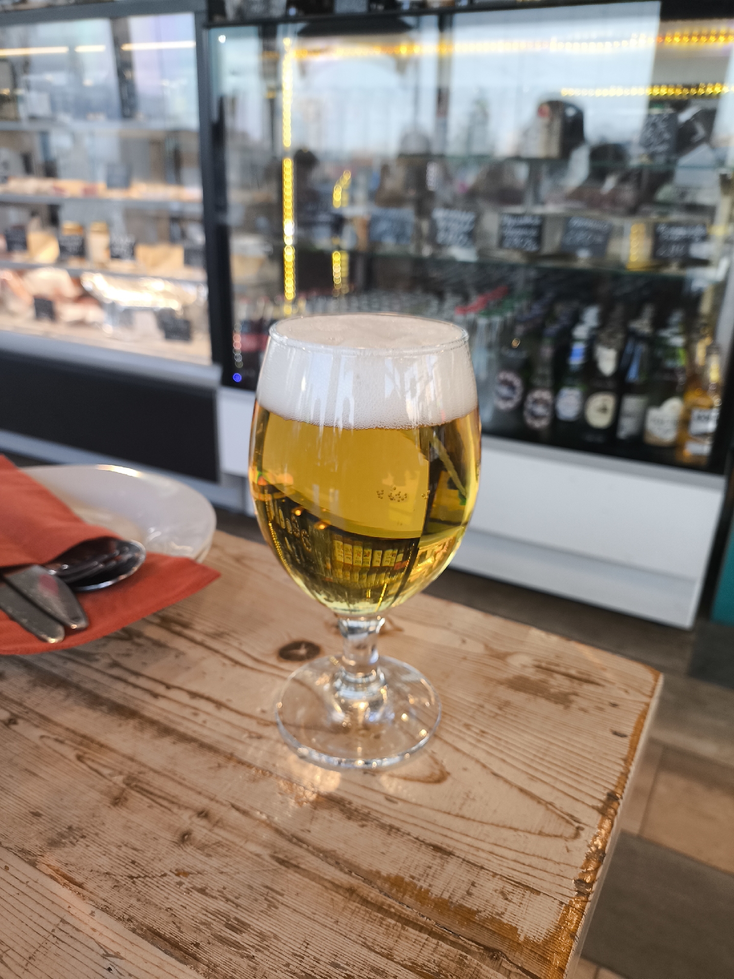 A full glass of golden beer with a thick white head sits on a wooden table. To the left, a plate with an orange napkin and silverware. Behind the beer, a refrigerated display case holds various items, including cheeses and bottled beverages. The location appears to be an establishment serving food and drinks, possibly in the city of Unknown, Unknown.
