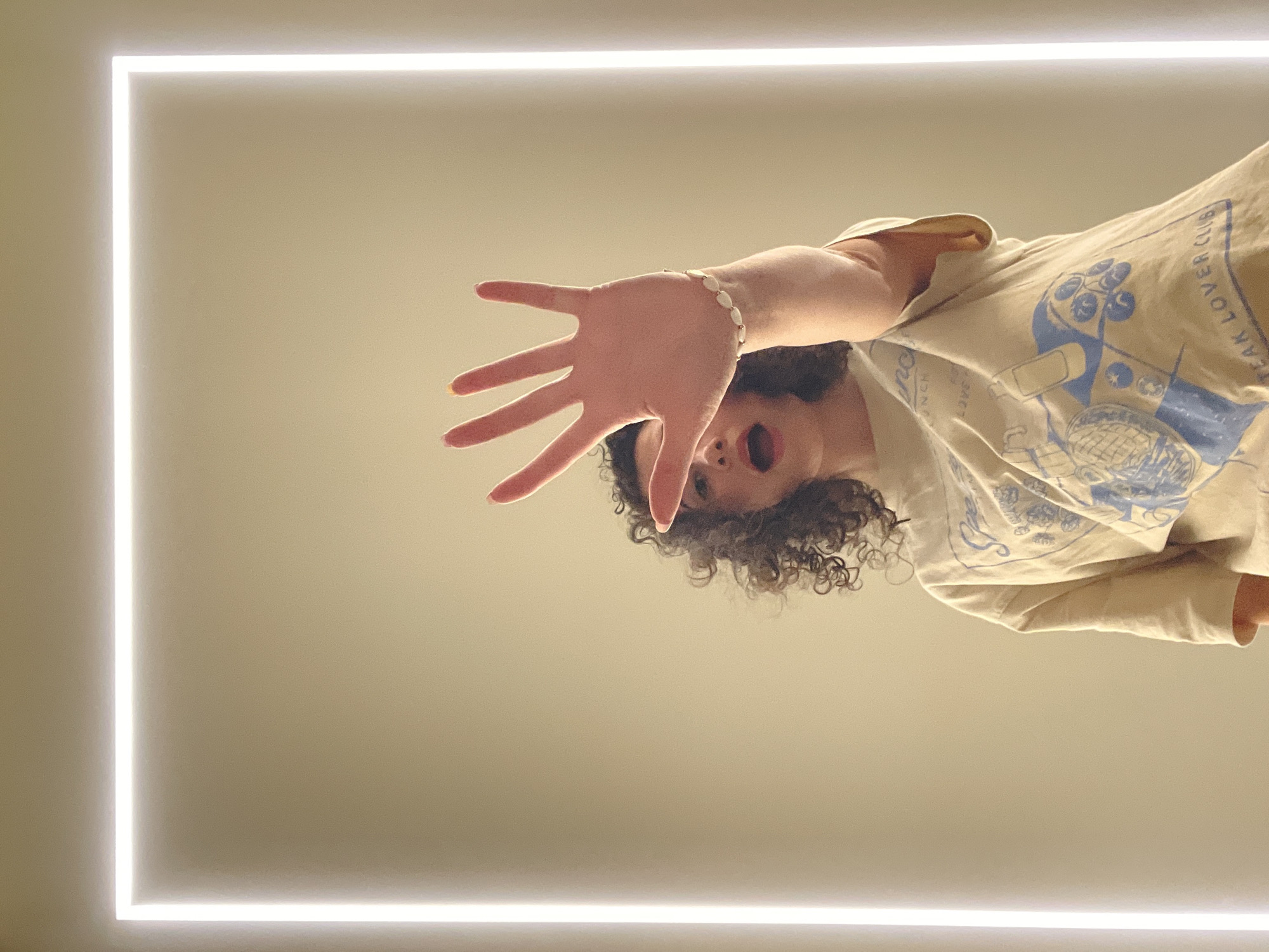 A person with curly hair and wearing a light-colored shirt with a blue design is positioned at the top-right of the frame, reaching out with their right hand towards the viewer. They have a bracelet on their wrist. The person's mouth is open, as if in surprise. The background is a light beige wall, framed by a rectangular LED light. There is no location information available in the image.