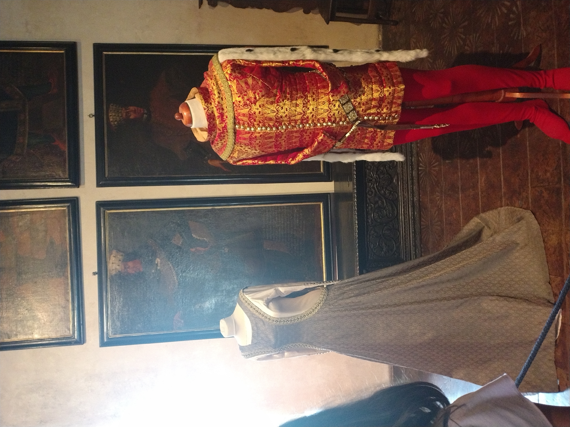 The image shows a museum display of historical clothing. On the left, a mannequin is dressed in a long, grey, patterned gown with a white underlayer and long sleeves. On the right, a headless mannequin wears a richly decorated red and gold tunic with a fur-lined collar and sleeves, red leggings, and a jeweled belt. Behind the mannequins are two large framed portraits depicting figures in historical attire. The room appears to have ornate wooden furniture and a tiled floor. The lighting is dim, with a bright light source coming from the left, casting shadows. The overall setting suggests a historical exhibition.