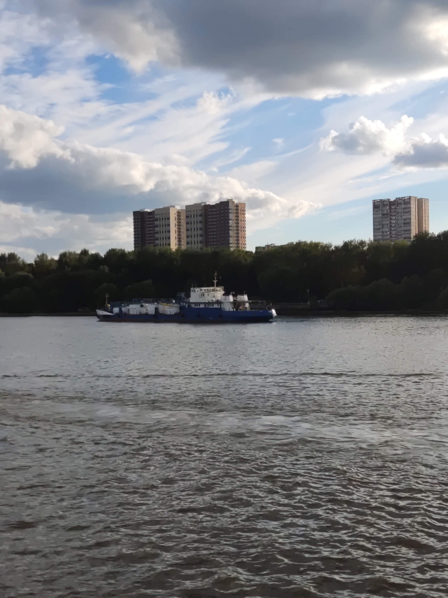 The image captures a wide shot of a body of water, likely a river or canal, in Khimki, Russia. In the distance, several tall, modern apartment buildings stand against a sky filled with a mix of white and dark clouds. A dense line of dark green trees separates the water from the buildings. A blue and white barge is visible on the water, moving away from the viewer. The water itself is a muted grey-green with visible ripples and small waves, suggesting movement. The overall scene appears to be an urban landscape with a focus on the interplay between nature and man-made structures.