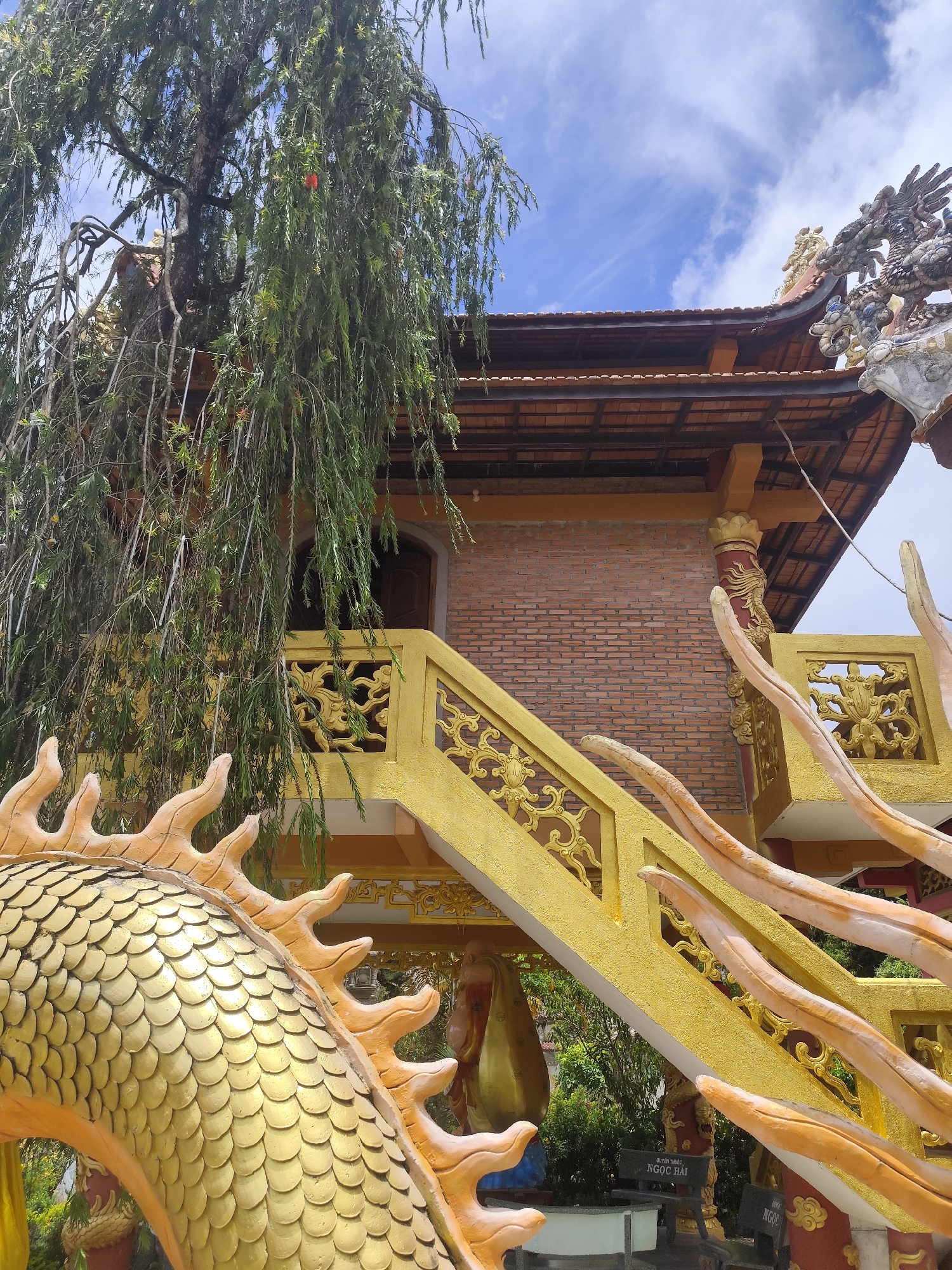 An outdoor, eye-level, medium shot captures a golden dragon sculpture in the foreground, positioned in front of a brick building with a tiled roof. The dragon's detailed scales are visible, along with its ornate horns and frills. A yellow, intricately designed staircase with dragon-like railing elements leads up to the building's entrance, which is partially obscured by a weeping willow tree with drooping branches. The building's brick facade is visible, and a decorative element with colorful ceramic pieces adorns the roofline. The sky above is a bright blue with scattered white clouds. In the lower portion of the image, a sign with Vietnamese text is affixed to a bench.
