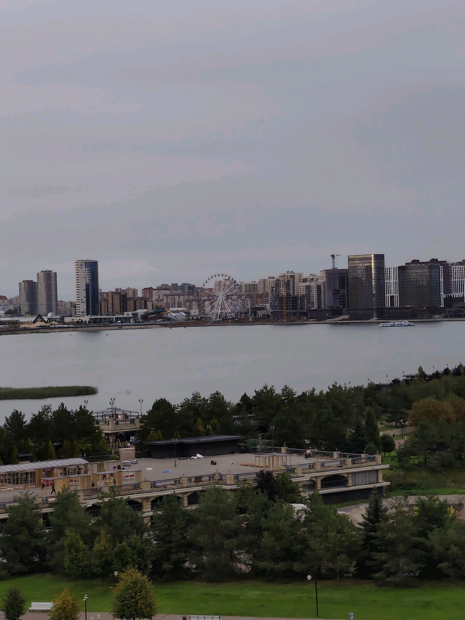 The image depicts a panoramic view of the Kazanka River in Kazan, Russia, with a modern cityscape lining its northern bank. In the foreground, lush green trees and a manicured lawn are visible, with a white bench placed on the grass. A paved walkway with lampposts runs through the park area.
The middle ground features a waterfront promenade with several structures. One building is multi-story with a colonnaded facade and a modern roof structure. Another building, appearing to be a park pavilion or restaurant, has a flat roof and glass enclosures. People can be seen walking on the promenade and near these structures. Dense trees with dark green foliage flank the promenade.
Across the Kazanka River, the cityscape of Kazan unfolds. Tall modern buildings with glass facades and varying architectural styles dominate the skyline. A Ferris wheel, a prominent landmark, is situated in the mid-distance, suggesting an entertainment area. The river itself is calm and reflects the overcast sky, with a pleasure boat cruising on its surface. The sky is heavily clouded, casting a soft, diffused light over the entire scene. The overall impression is one of a contemporary urban waterfront with recreational amenities and a backdrop of modern architecture.