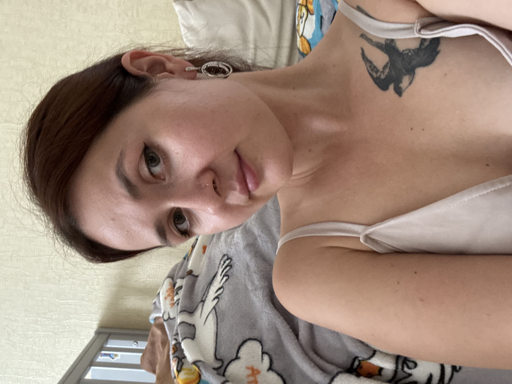 An individual is captured in a close-up selfie, their head and upper torso filling the frame. They are lying down, with a grey blanket featuring cartoon characters partially visible on the left and a white pillow behind them on the right. The individual has brown hair pulled back, a piercing on their nose, and is wearing a light-colored tank top. A tattoo of a bird is visible on their chest. They are also wearing an earring in their right ear. The background appears to be a wall with textured wallpaper and a portion of a metal bed frame. There is no information in the image to indicate a location in Naberezhnye Chelny, Russia.