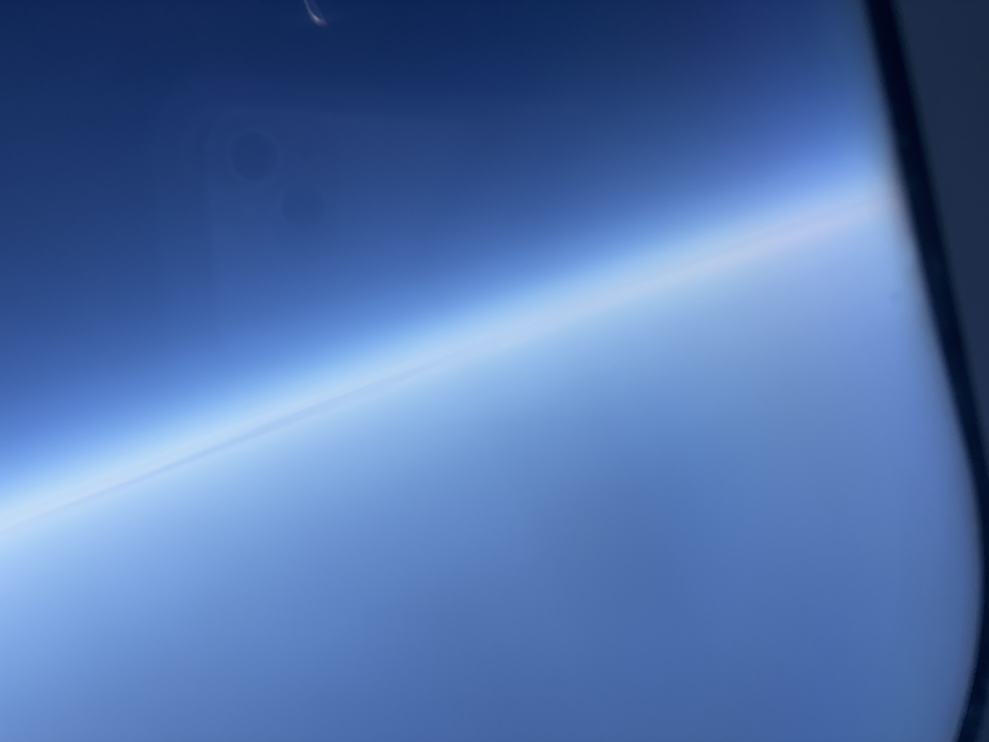 The image is a blurred, high-angle shot of the Earth's atmosphere seen from above. The sky transitions from a deep blue at the top to a lighter blue near the horizon line, which is softly illuminated by sunlight. There are faint, circular smudges on the surface of what appears to be a window, and a white streak of light is visible at the top left, possibly from movement or a reflection. The right side of the image shows a dark, blurred edge, suggesting the boundary of the window or viewing frame. There are no people, subjects, or identifiable objects in the scene. The location is unknown.