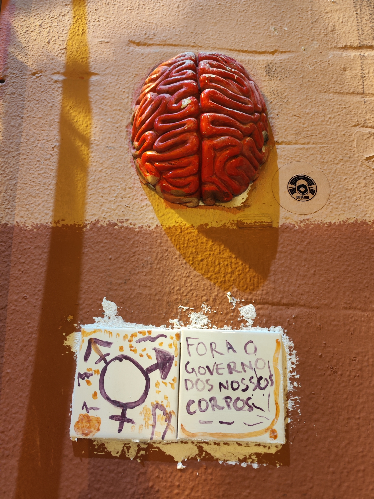 A close-up shot shows a section of a weathered, two-toned wall, possibly an exterior or interior surface. The upper part of the wall is painted a mottled yellowish-beige color, transitioning to a darker, reddish-brown lower section. Affixed to the upper part of the wall is a red, anatomically stylized brain sculpture. Below the sculpture, slightly above the color transition, are two ceramic tiles. The left tile features a hand-drawn symbol representing transgender identity in purple paint. The right tile contains Portuguese writing, also in purple paint: "Fora o Governo dos Nossos Corpos!", translating to "Government out of our bodies!" The tiles appear to be secured to the wall with a white, plaster-like substance, that is also visible around and below the tiles and sculpture, indicating repair or modification of the wall. A circular sticker is affixed to the wall to the right of the brain, depicting a stylized logo with the word "Outlaw" visible at the bottom. A shadow, possibly from a nearby object or light source, is cast across the upper section of the wall, adding to the textured appearance.
The political graffiti and artwork suggest the image was taken in a location where activism is prevalent. Lisbon, Portugal, is known for its street art and political expression.