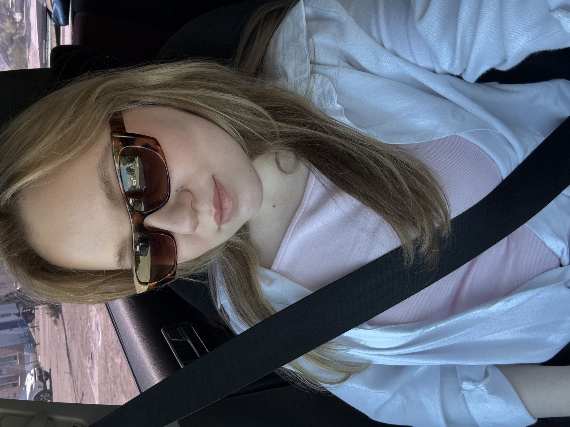 A young Caucasian woman with blonde hair, wearing sunglasses, a pink top, and an open white button-down shirt, is seated in the passenger seat of a car. She is wearing a black seatbelt. The interior of the car is visible, with a black door panel and seat. Outside the car, through the window, a street scene in Chaikovsky, Russia, is depicted. A white car is parked on a dirt road, with buildings and trees in the background. The sun is shining, creating a bright outdoor environment.