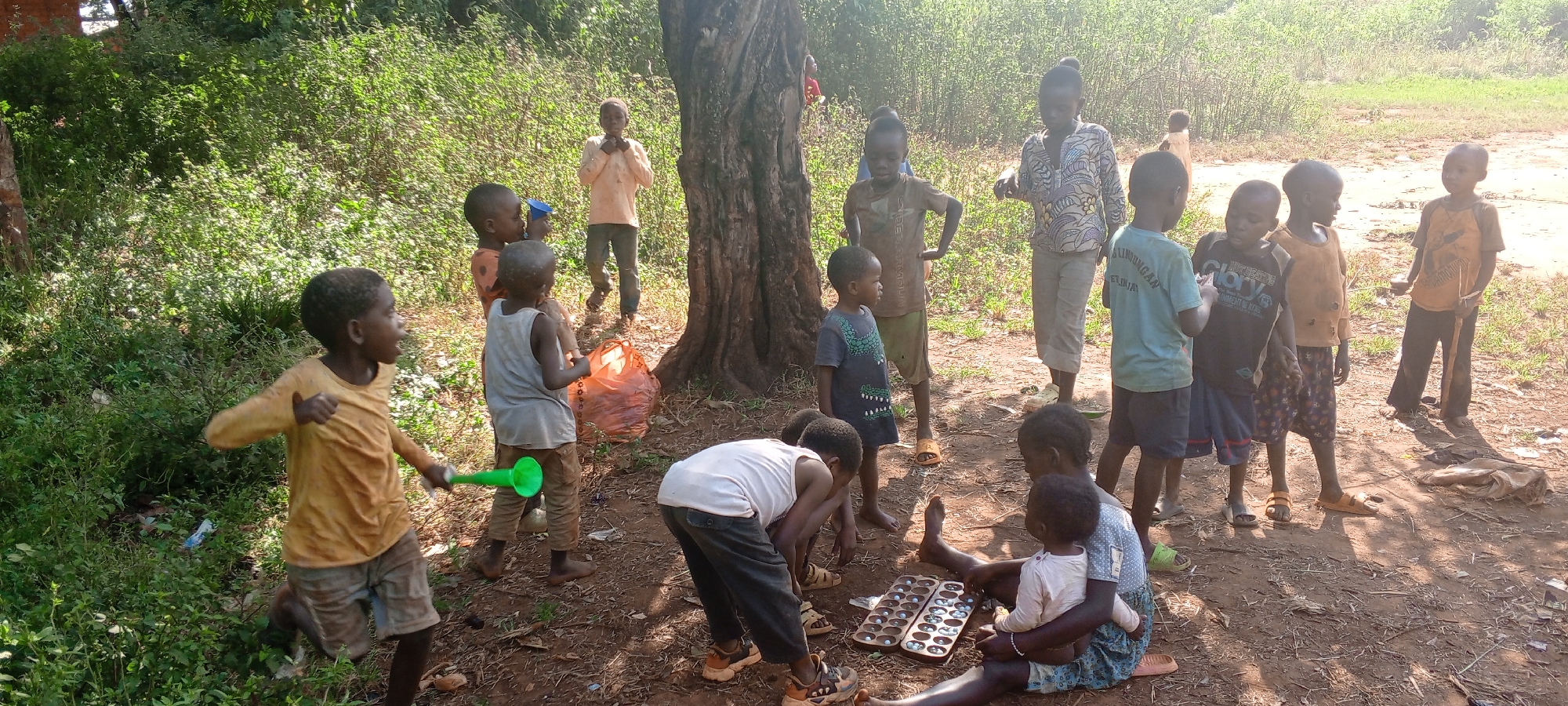 The image shows a group of children and at least one adult in a grassy area in Ngalula, Democratic Republic of the Congo. A tree dominates the center of the frame. Several children are standing and interacting near the tree, while one child runs in the foreground, holding a green object. Another group sits on the ground, gathered around a board game. The adult figure stands among the children, wearing a patterned shirt and pants. The environment includes dense vegetation and a dirt ground.