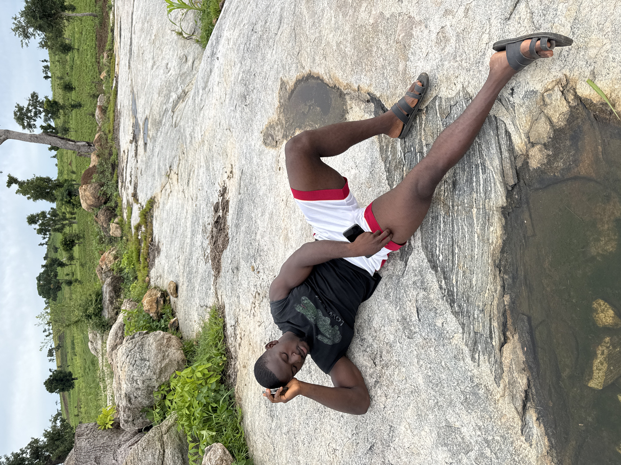 A Black man is lying on his side on a large, flat, grey rock surface. He is wearing a black sleeveless t-shirt with a green alligator logo and white shorts with red trim. His left arm is bent at the elbow, supporting his head as he looks down with his eyes closed. His right leg is bent at the knee and extended outwards, and his left leg is bent at the knee and tucked in. He is wearing dark sandals on both feet. The rock surface has some damp patches and small puddles. In the background, there is a gently sloping, green, grassy hill dotted with scattered trees. The sky is overcast. The scene appears to be an outdoor natural area, possibly in or near Akwanga, Nigeria.