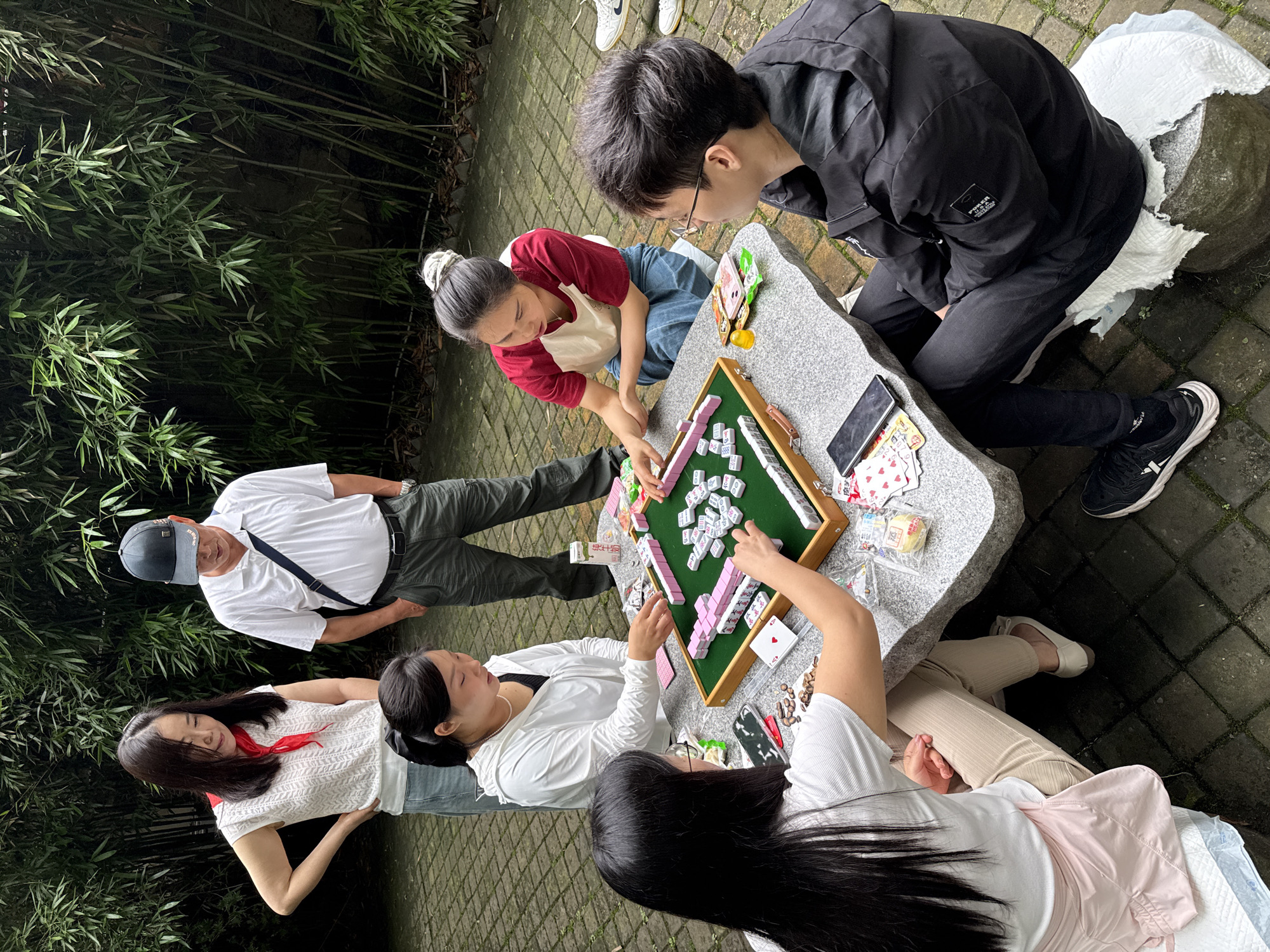 In Yuzhong District, China, a group of six people are gathered around a stone table on a paved surface outdoors. They are playing mahjong, as evidenced by the mahjong tiles and board on the table. A person wearing glasses and a black jacket is sitting and observing the game. A woman in a red and white shirt is reaching towards the tiles. Another woman wearing a white jacket is participating in the game. Two other women are also involved in the game, and an older man in a white shirt and cap is standing nearby watching. A smartphone and other small items are also present on the table. Bamboo stalks are visible in the background.