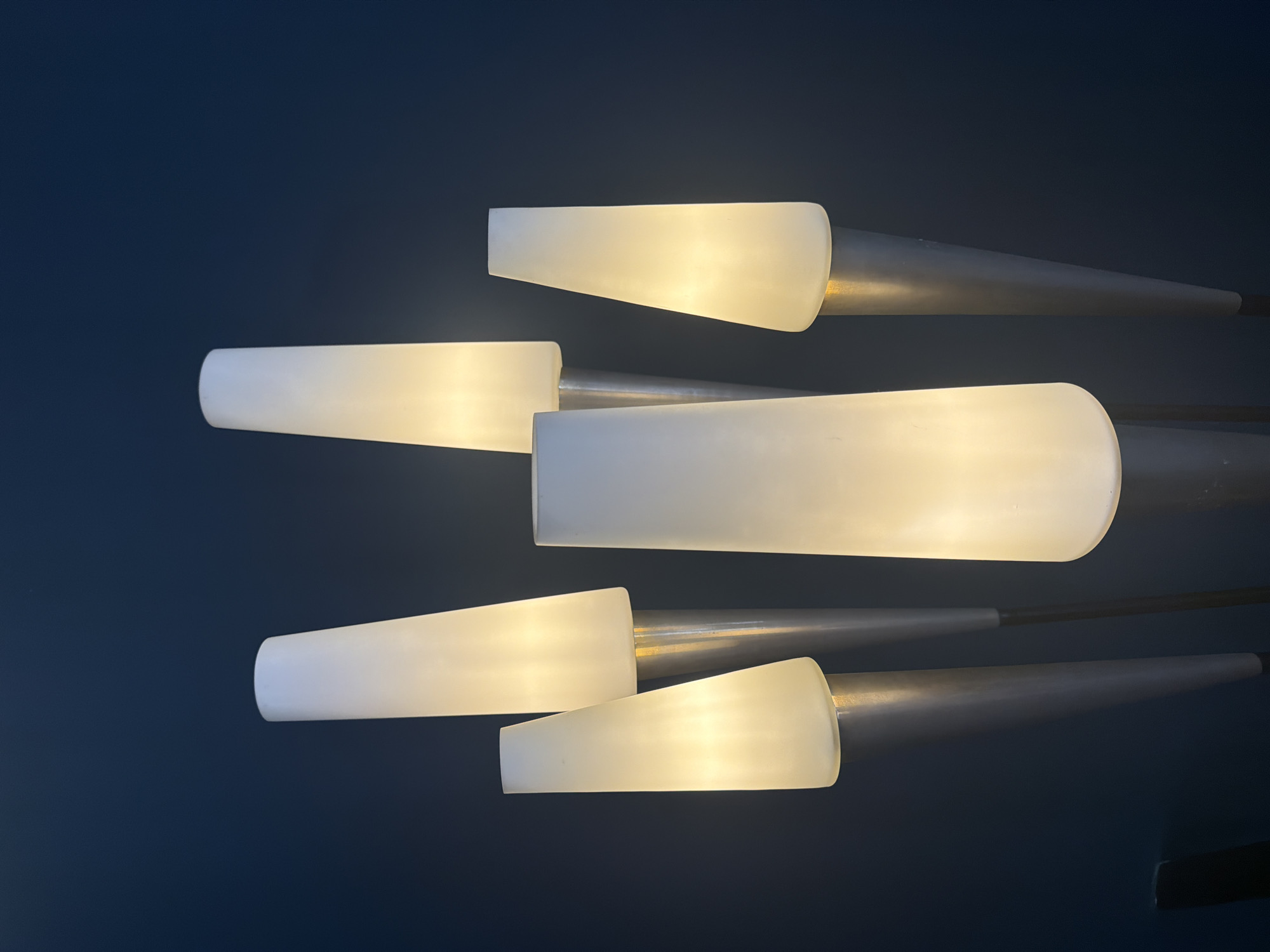The image depicts a portion of a light fixture against a dark blue background, likely a ceiling or wall. The fixture consists of multiple white, opaque, elongated shades, illuminated from within, attached to what appear to be metallic rods or arms. The shapes of the shades vary slightly, some are more cylindrical while others taper. The rods extend outward from the fixture, and the overall arrangement is somewhat abstract. The photo was taken at the Hotel Plaza Athenee in Paris.